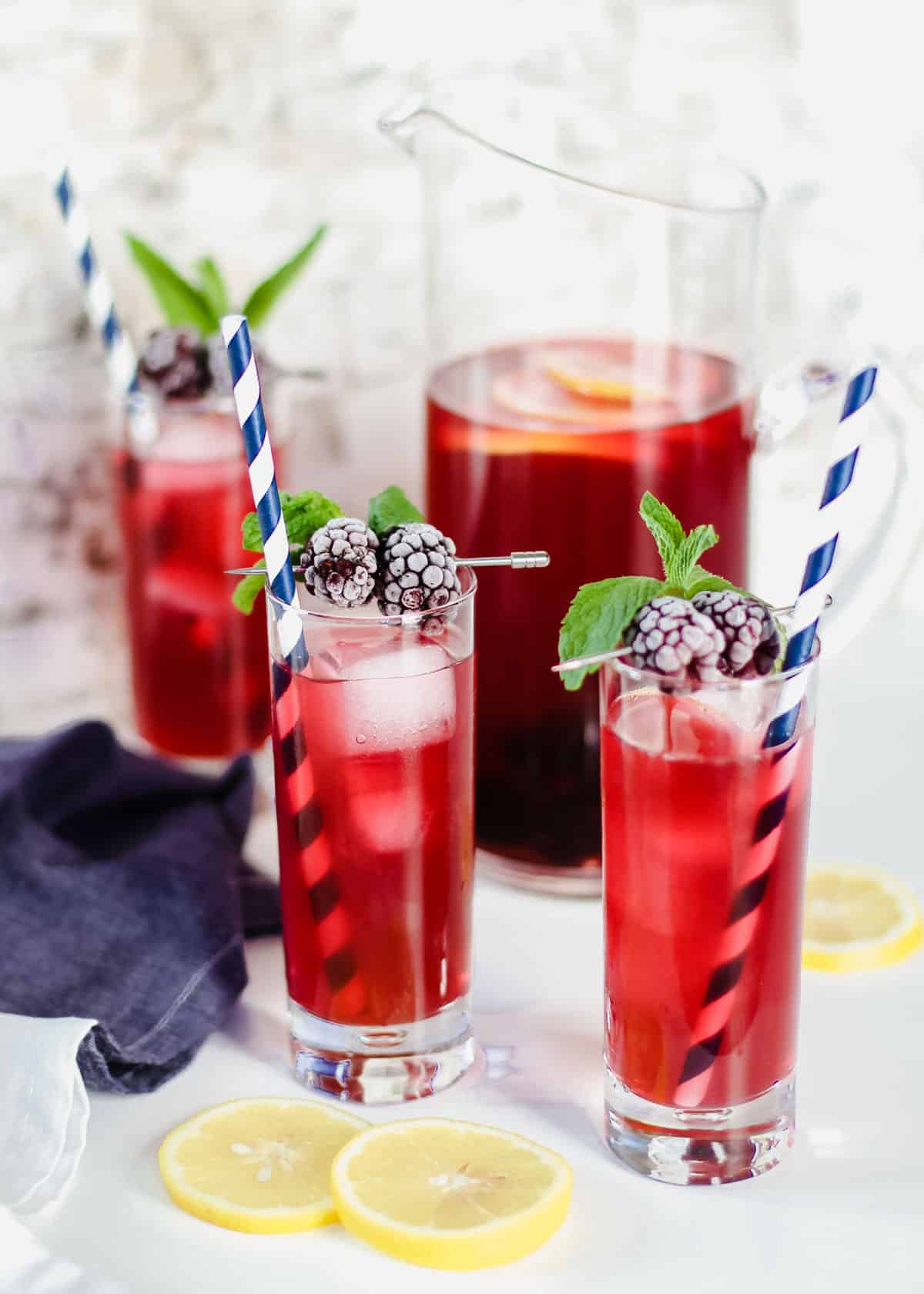 three tall glasses filled with  red drink on ice garnished with blackberries and mint, with pitcher in background.