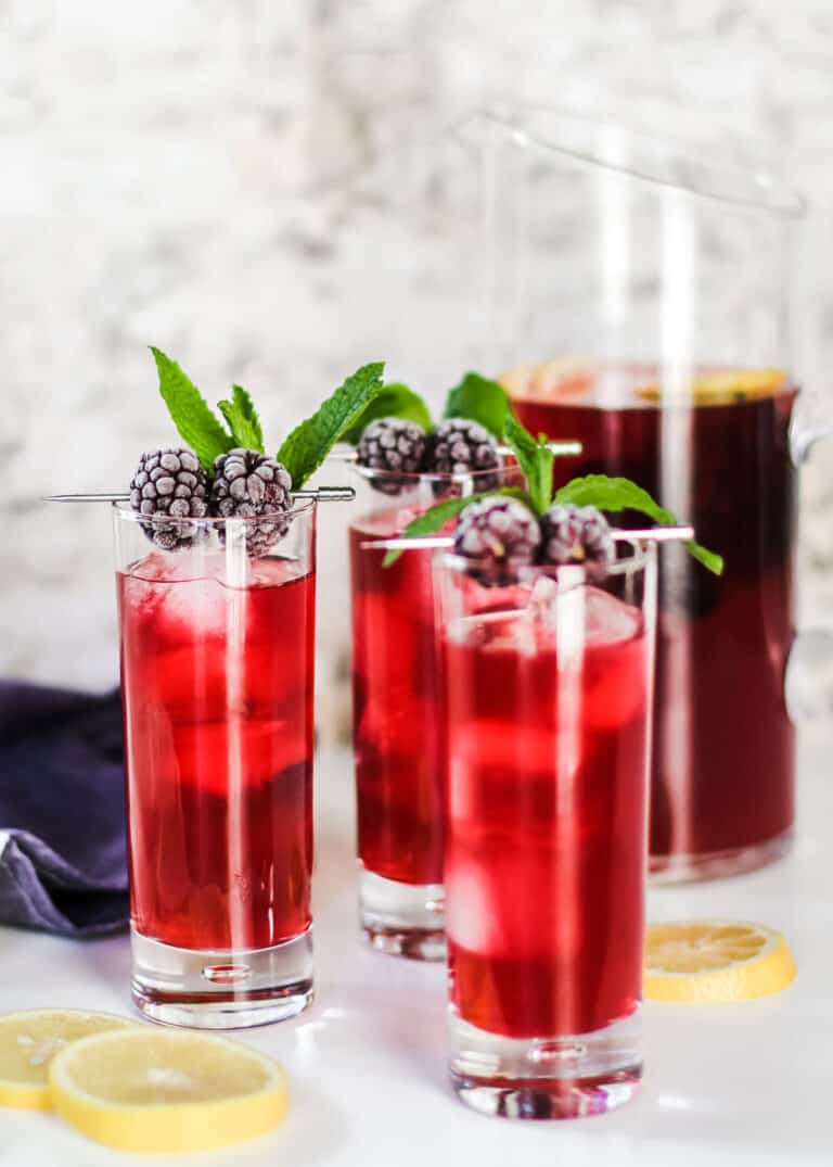 three tall glasses filled with red drink on ice garnished with blackberries and mint, with pitcher in background.