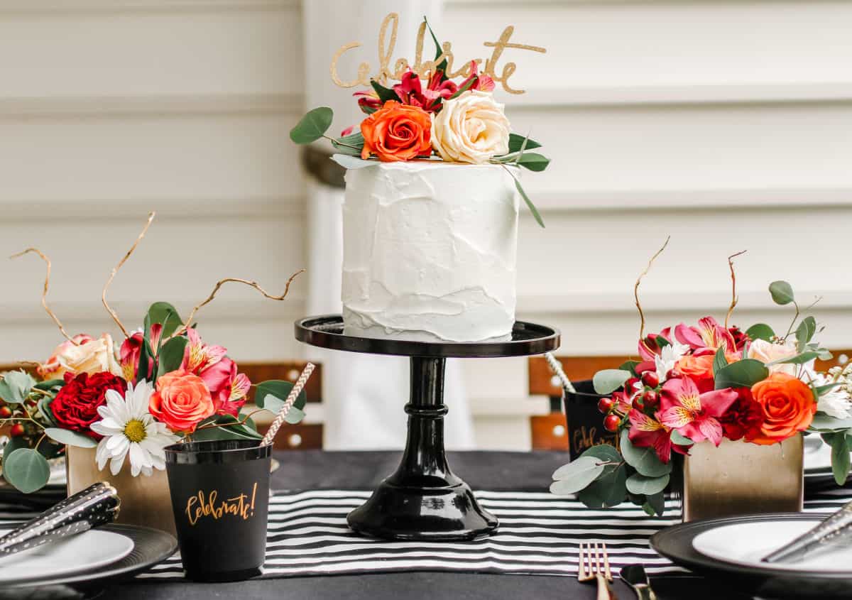 tablescape centerpiece of two colorful flower arrangements and white cake on cake stand.