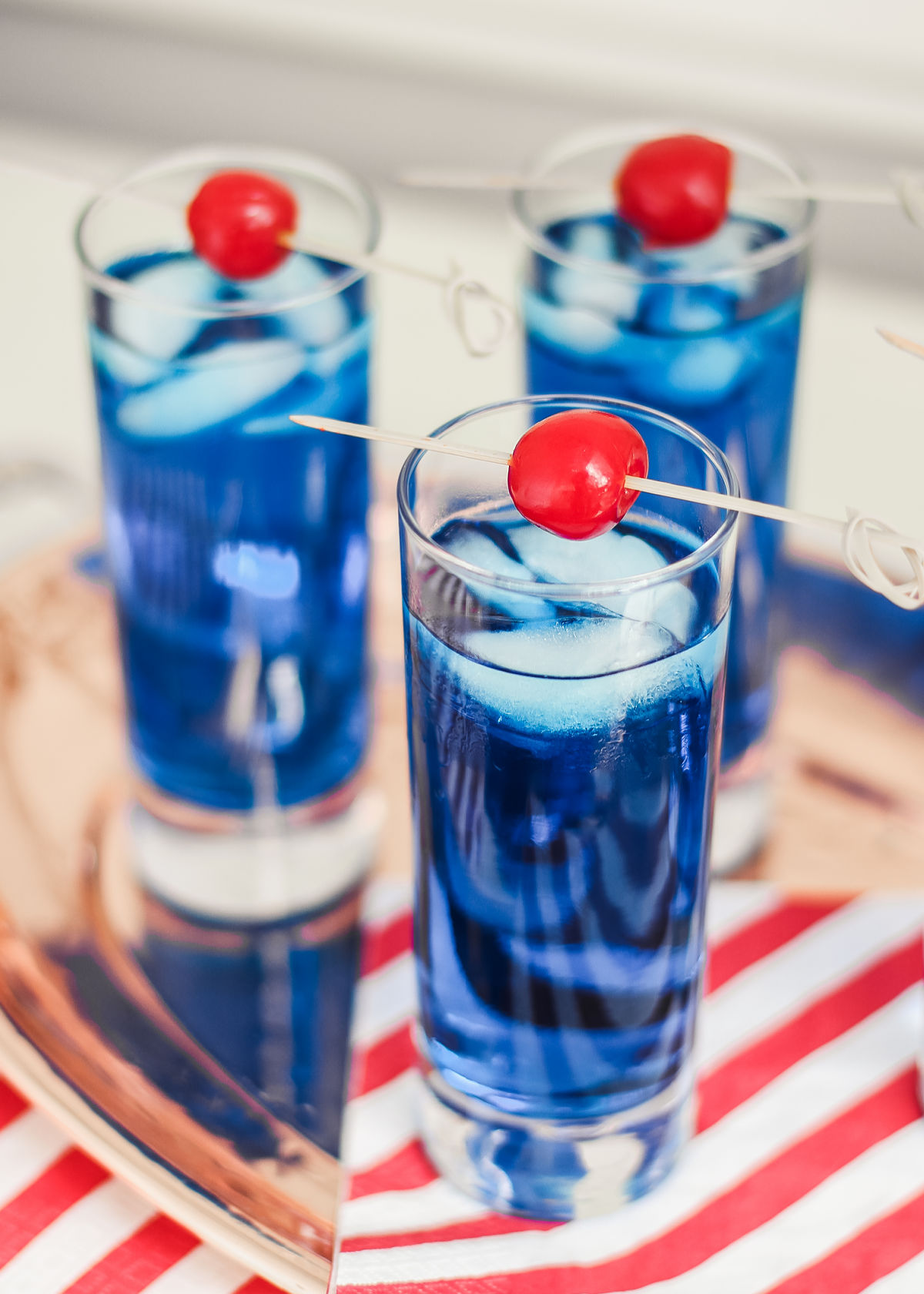3 glasses with blue drink and cherry garnish on tray.