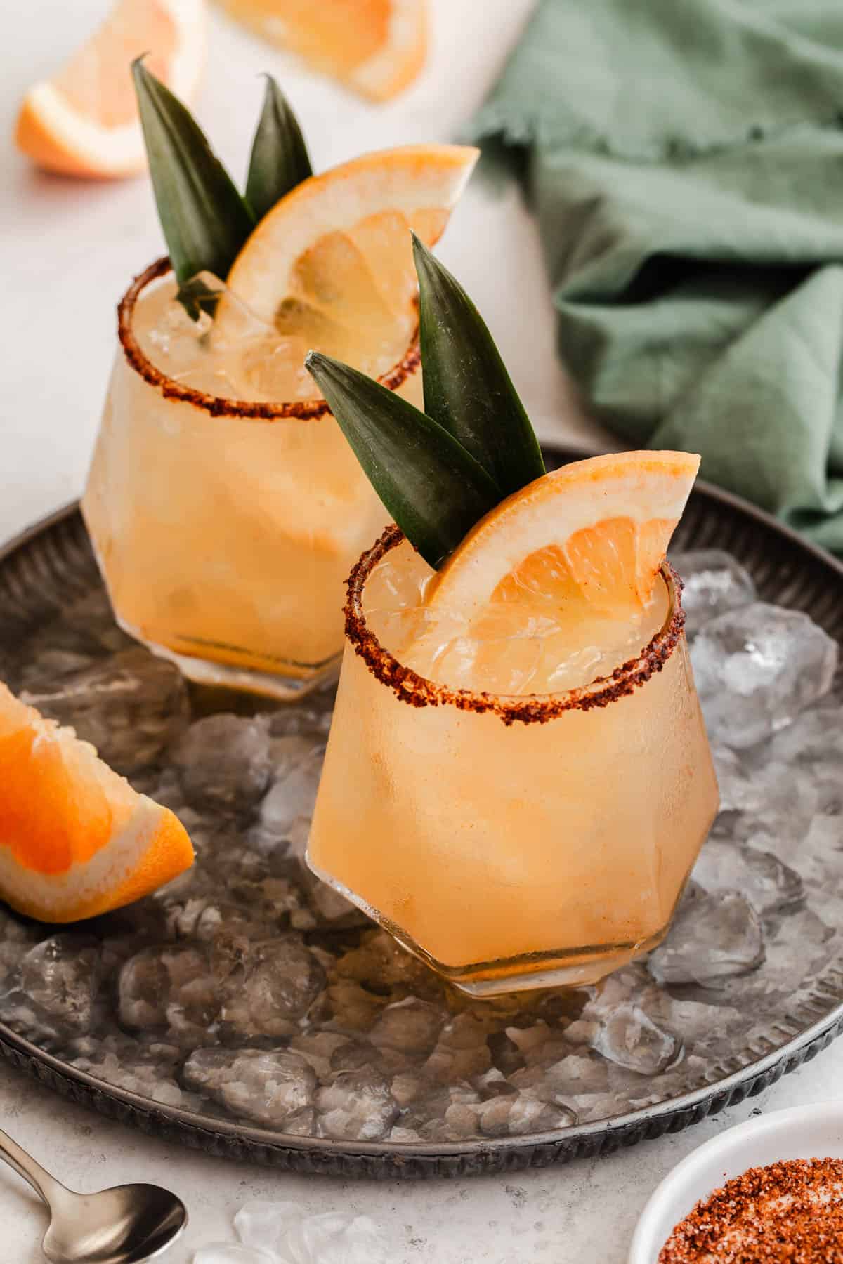 two glasses with peach colored drinks garnished with grapefruit slices and pineapple leaves, on gray tray.