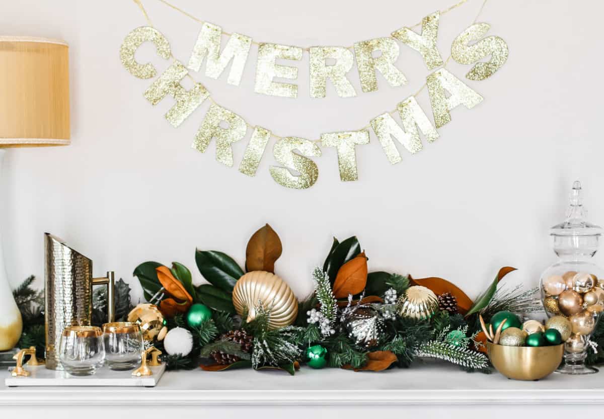 white buffet table with greenery runner and gold ornaments, with Merry Christmas banner hanging on wall above.