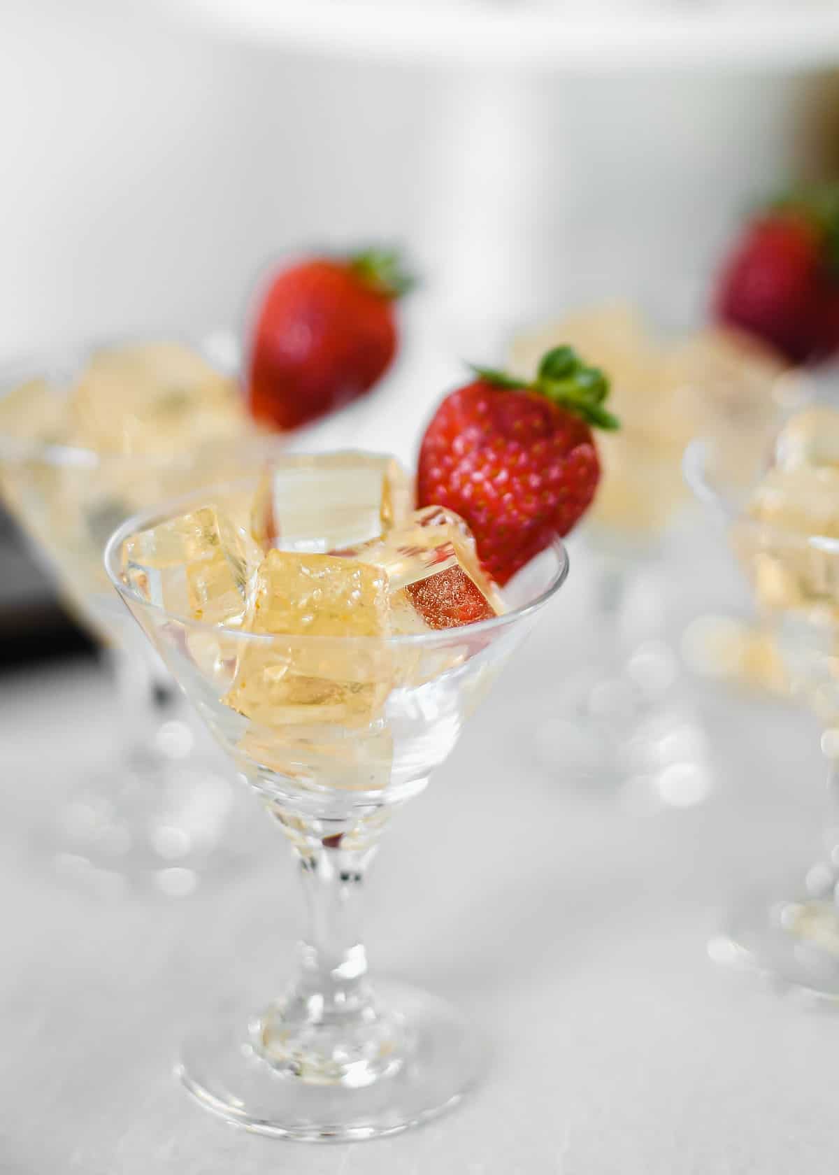 mini martini glasses with strawberry garnish on the side, filled with golden colored jello squares.