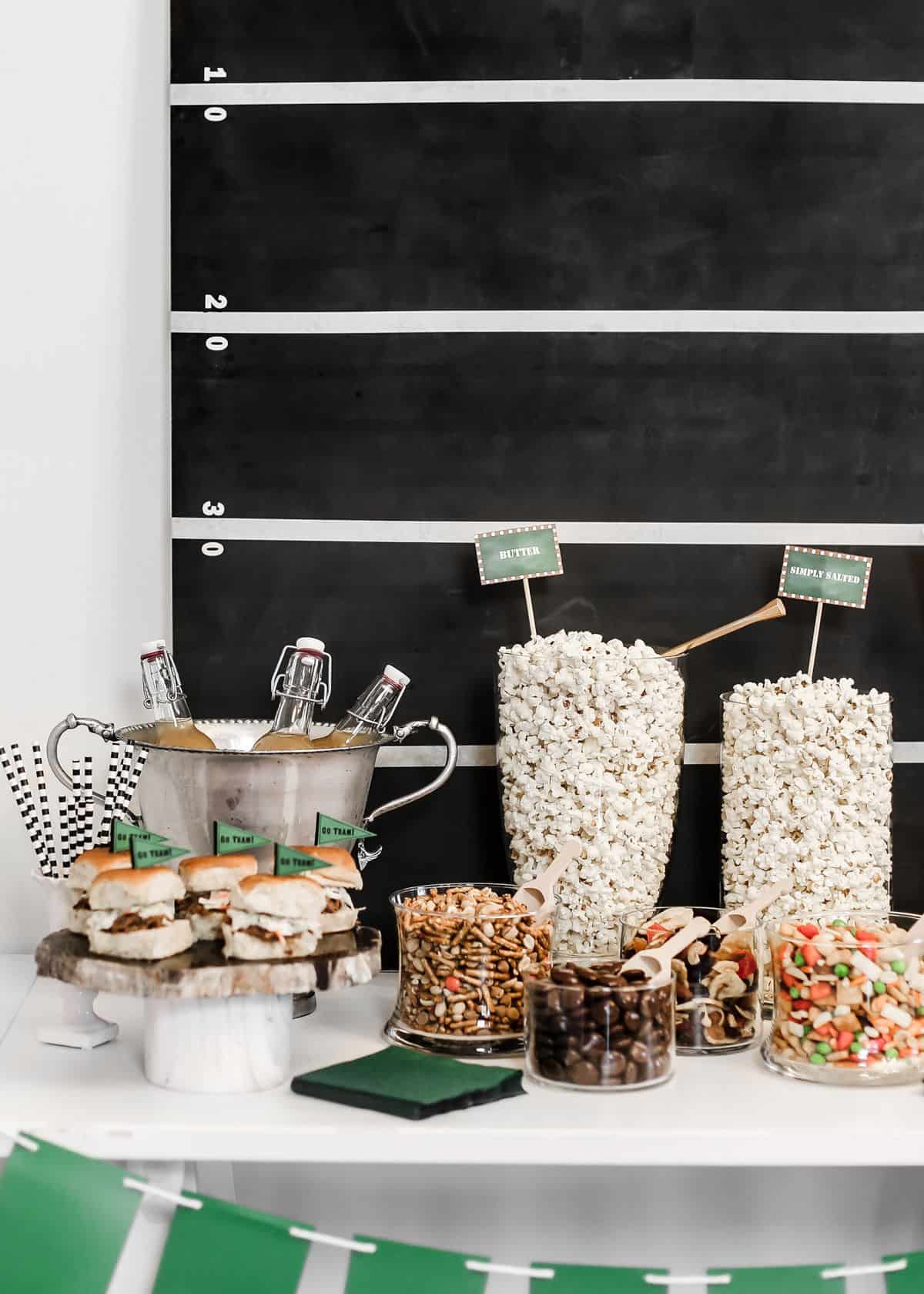 snack buffet with mini sandwiches, popcorn in jars, and drink bottles in silver urn.