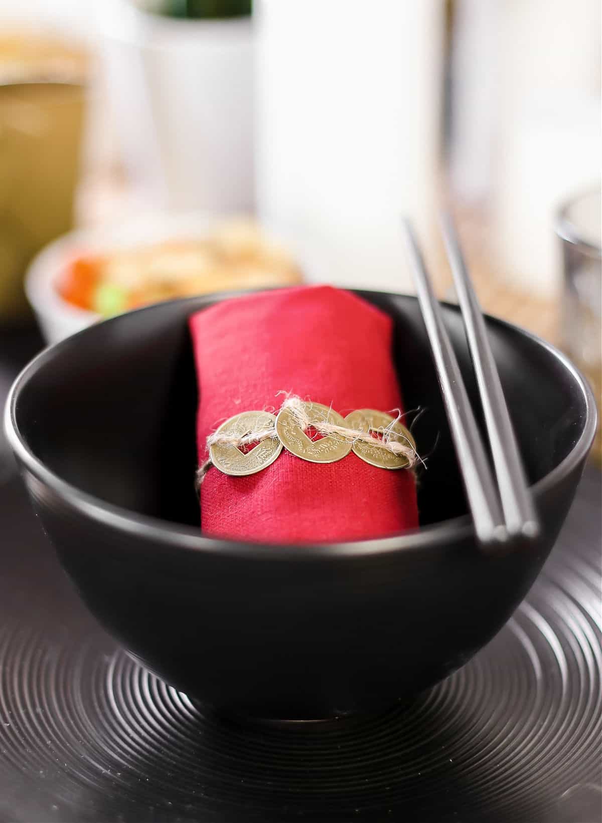 red napkin rolled up and ties with chinese good luck coins, sitting in a black bowl