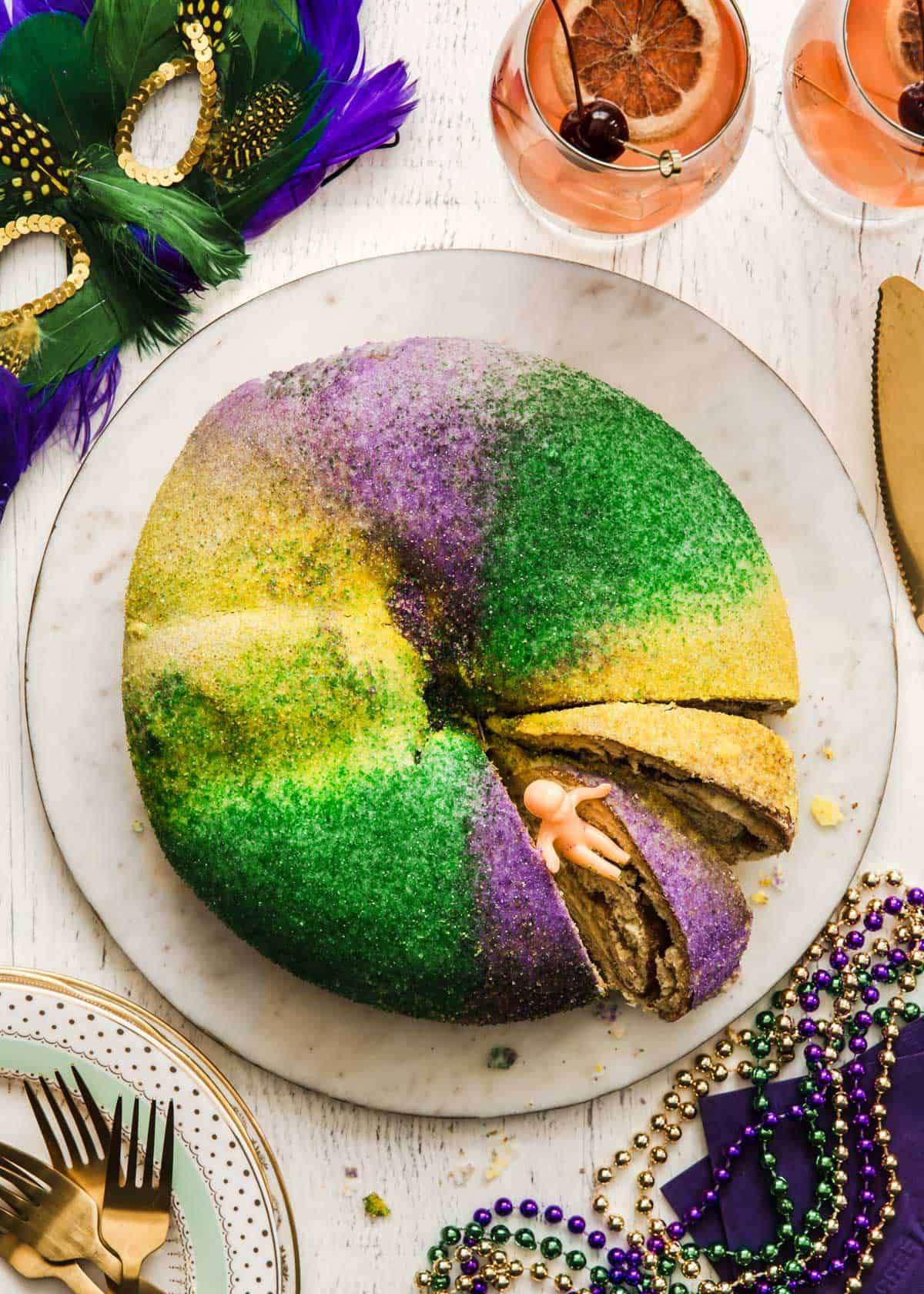 Mardi Gras King cake on white platter surrounded by mask, beads, and hurricane drinks.