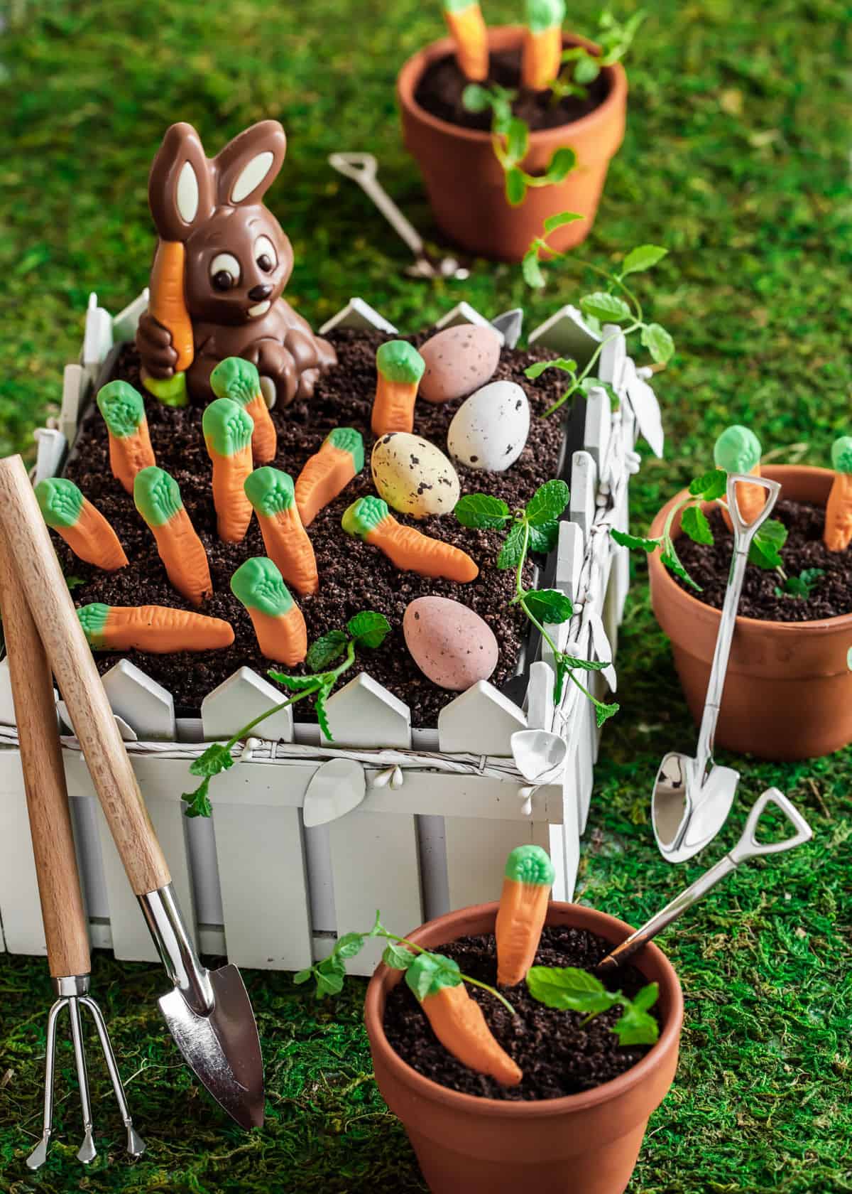 cute Easter garden dirt cake with candy carrots and chocolate bunny, in fence shaped serving vessel, on moss grass runner.