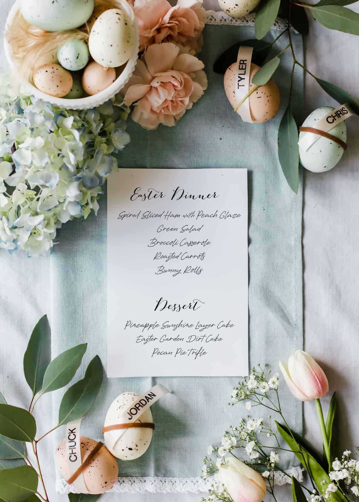 printed menu card sitting on a blue napkin surrounded by Easter eggs and fresh spring flowers.