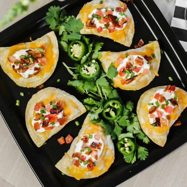 black square plate with pierogies topped with cheese, sour cream, tomatoes and bacon.