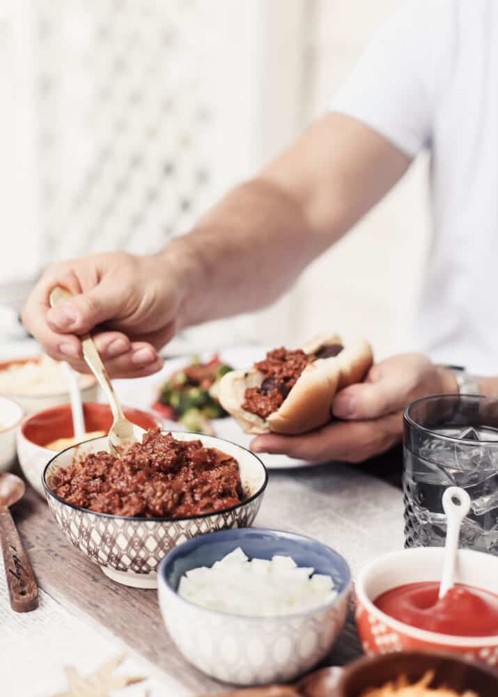 table with man holding a hot dog surrounded by toppings, scooping out chili from a bowl.