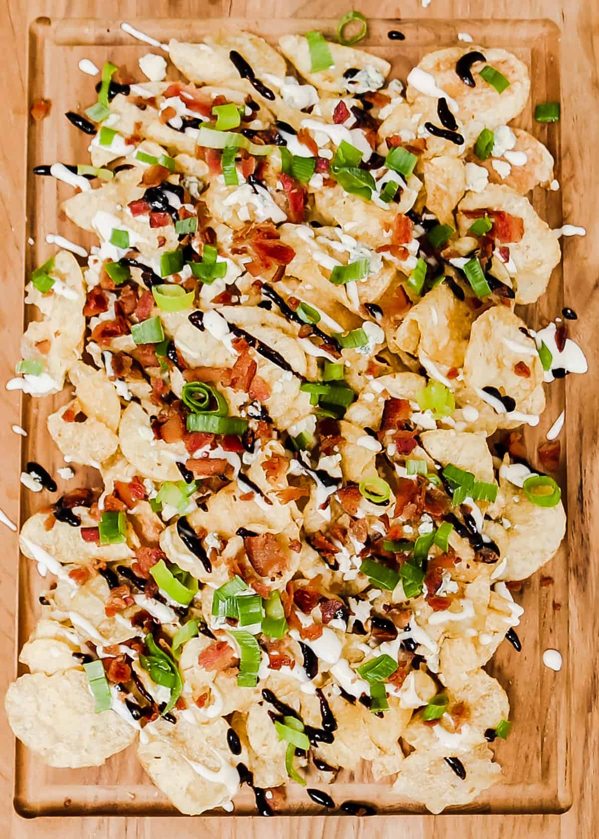 piles of potato chips topped with bacon, green onion, white sauce and balsamic glaze, on wood serving board.
