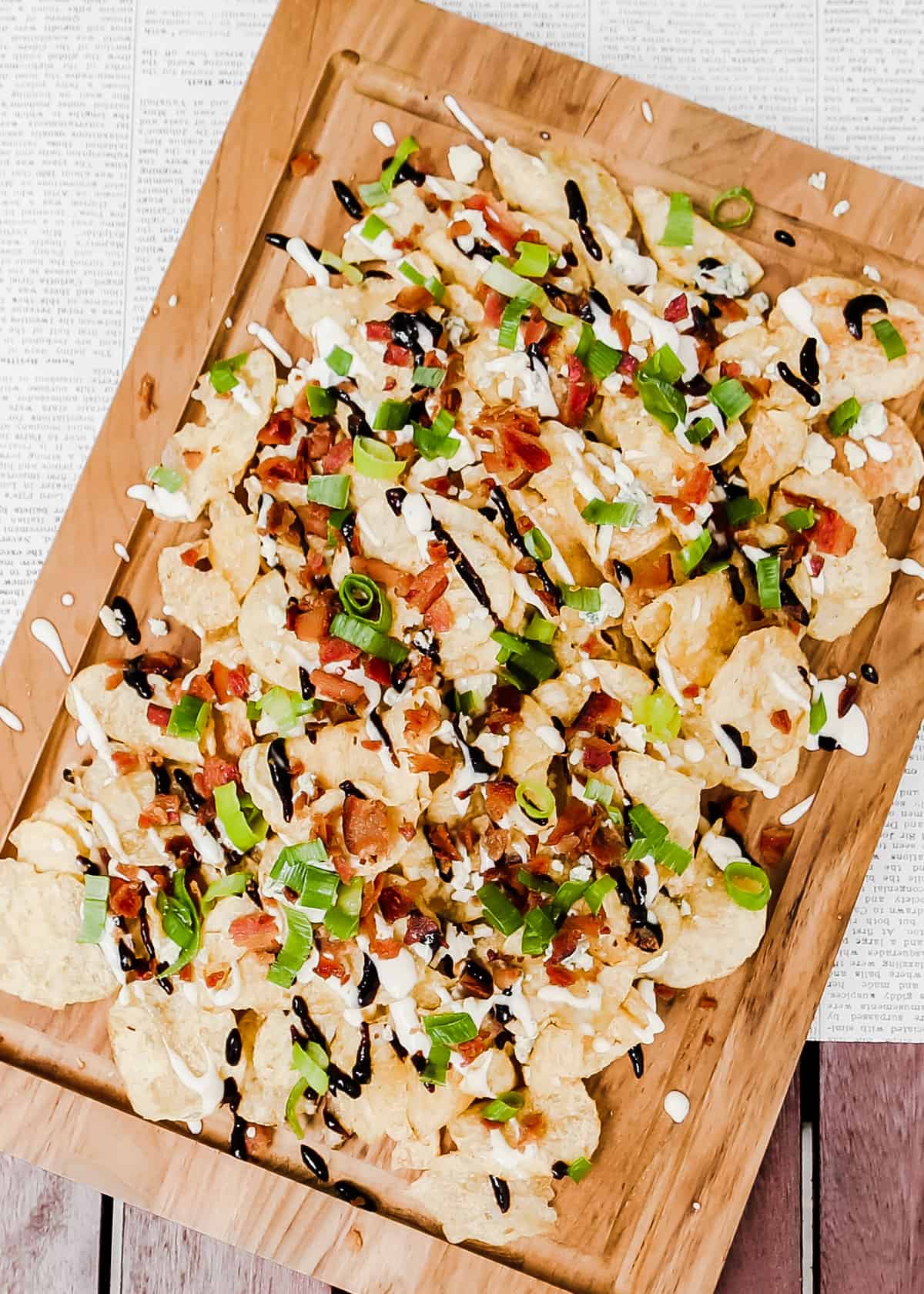 piles of potato chips topped with bacon, green onion, white sauce and balsamic glaze, on wood serving board.