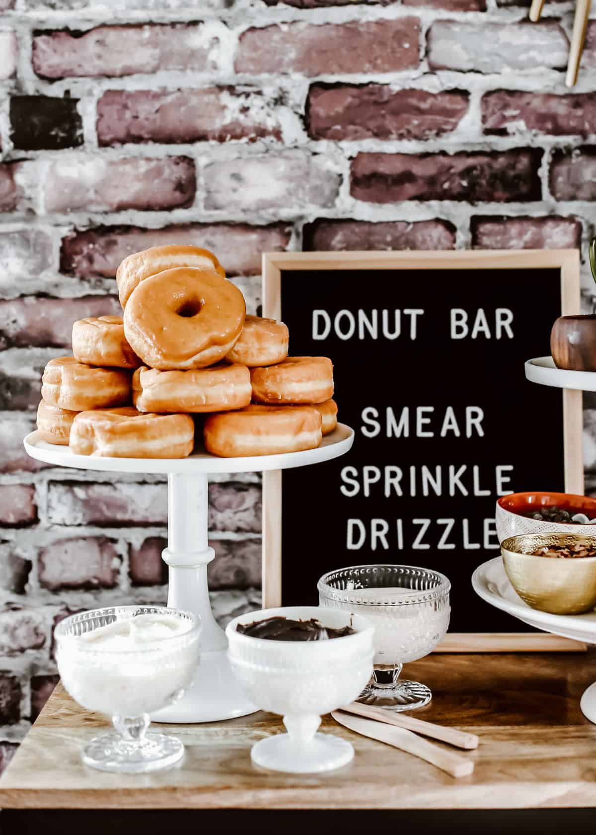glazed donuts stacked on white cake stand surrounded by toppings in bowls, on table with brick wall background.