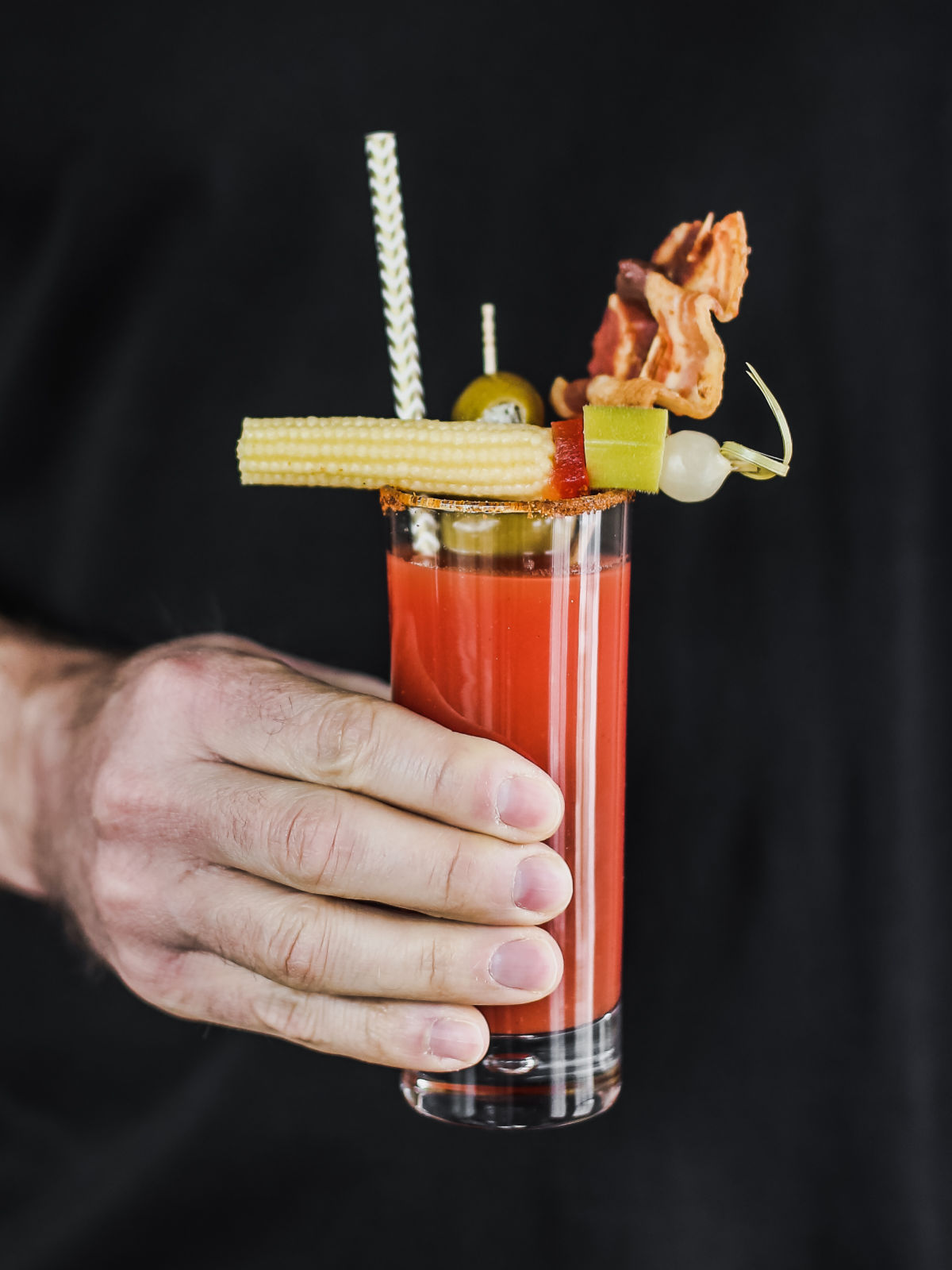 hand holding bloody mary in glass with bacon and baby corn garnish.