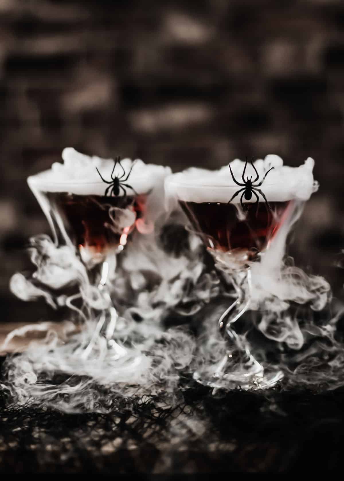 2 martini glasses with dark drinks and dry ice smoke, and spider on side of glass.