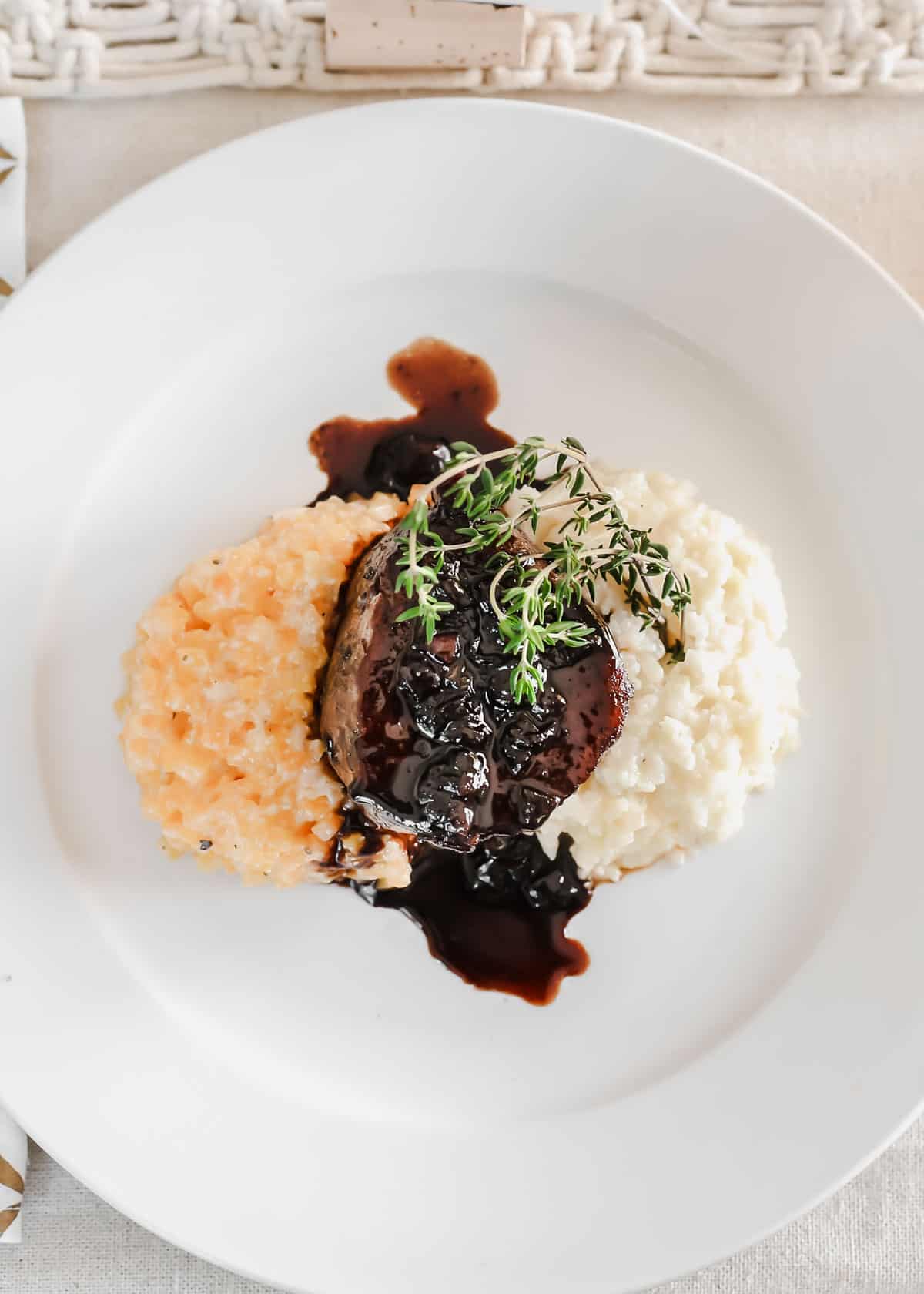 filet mignon topped with glaze, served on a bed of risotto and riced cauliflower on white plate.