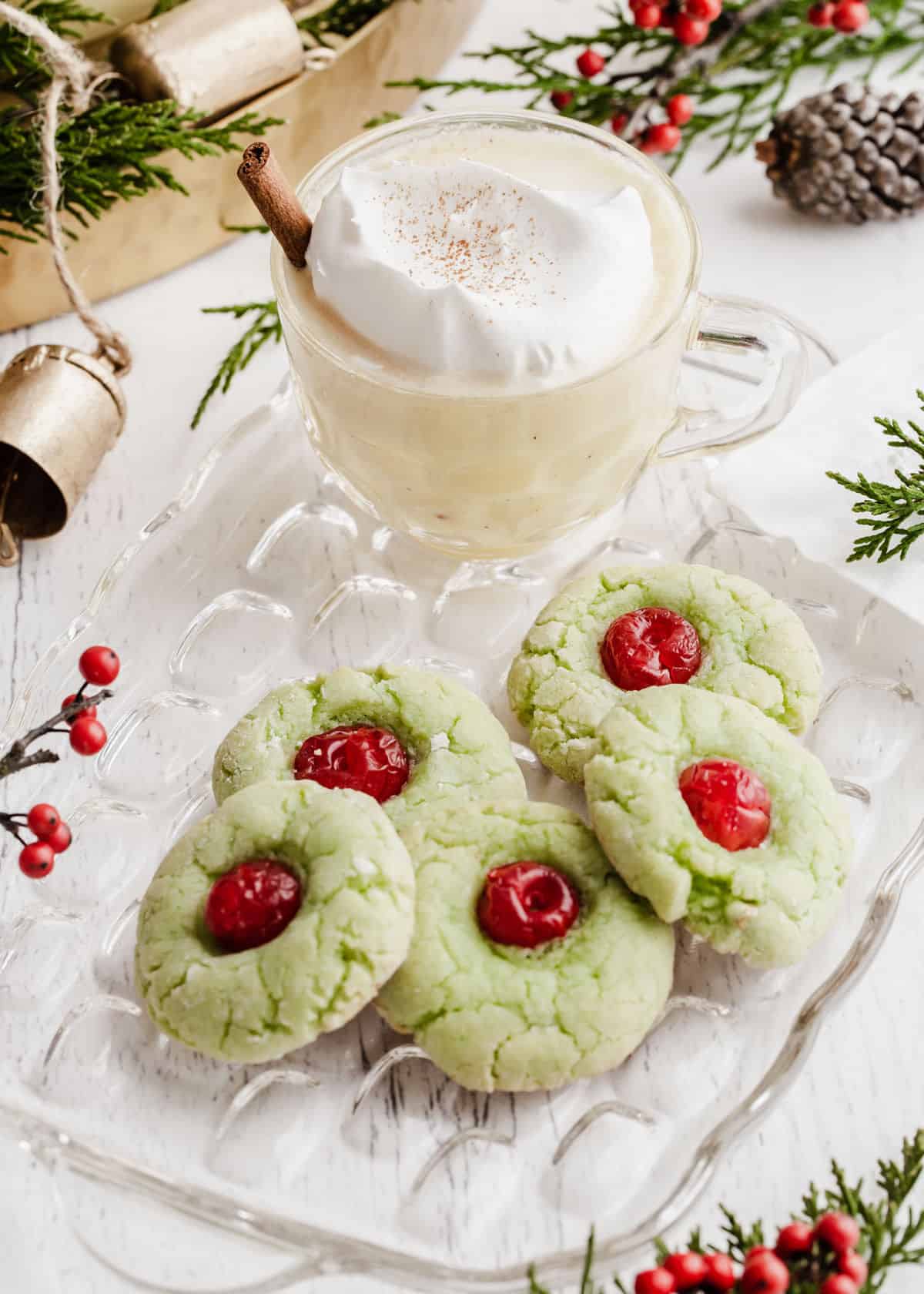 green cookies with red cherry in the middle, on glass plate with cup of eggnog.
