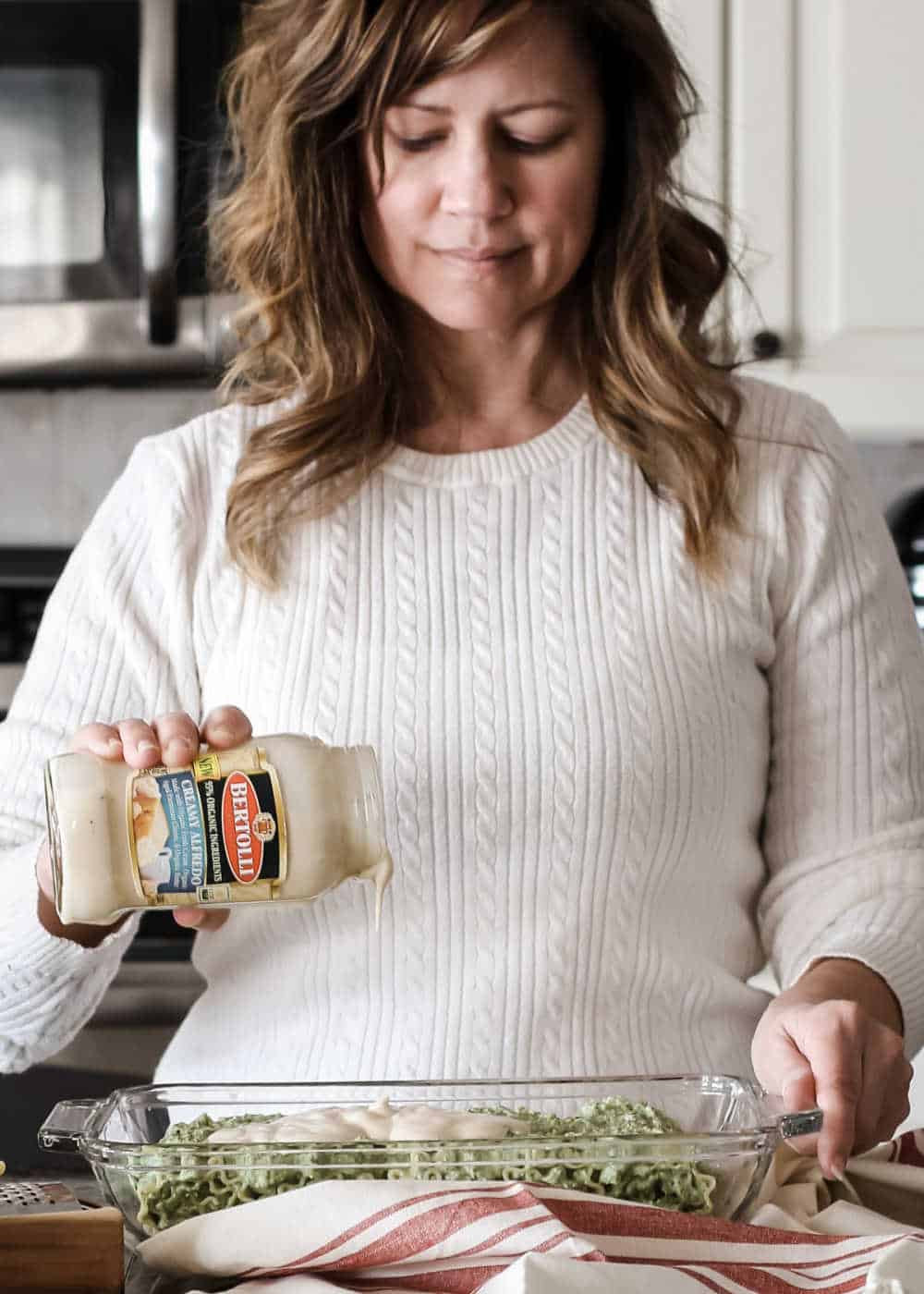 woman in kitchen pouring alfredo sauce over food in casserole dish.