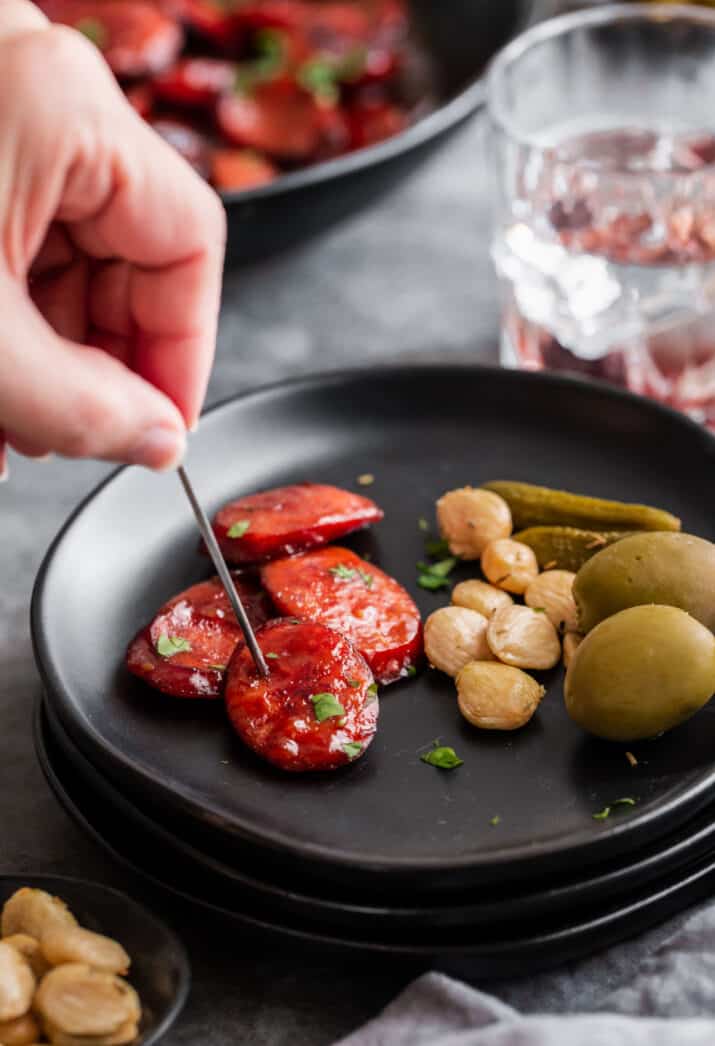 black appetizer plate with saucy sliced kielbasa and hand using a pick to grab it.