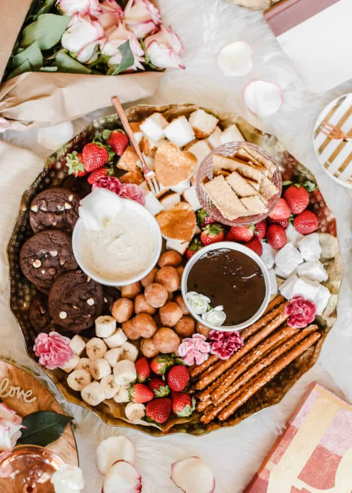 overhead view of gold round tray filled with 2 bowls of sweet fondue and dippers like strawberries, pretzel rods, bananas, cookies, marshmallows, graham crackers and donut holes.