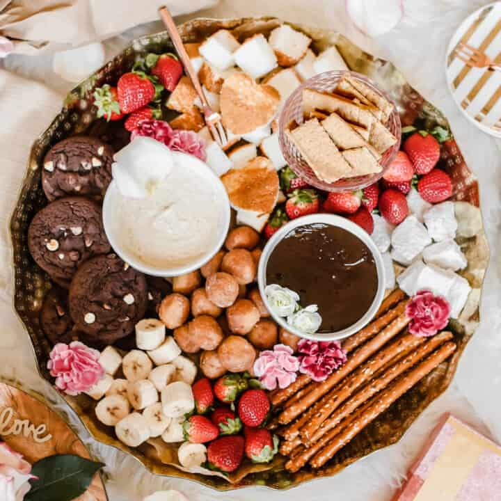gold round tray filled with 2 bowls of sweet fondue and dippers like strawberries, pretzel rods, bananas, cookies, marshmallows, graham crackers and donut holes.