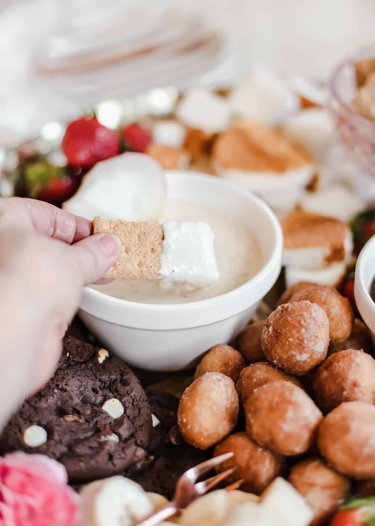 hand dipping graham cracker into white dessert fondue dip.