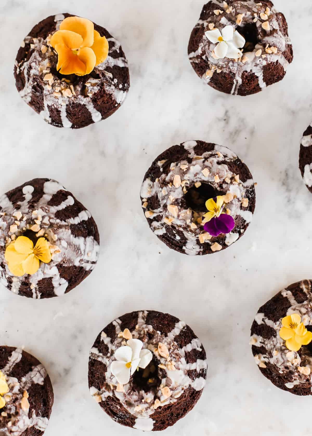 rows of mini chocolate cakes with flowers garnish, on marble table, overhead view.