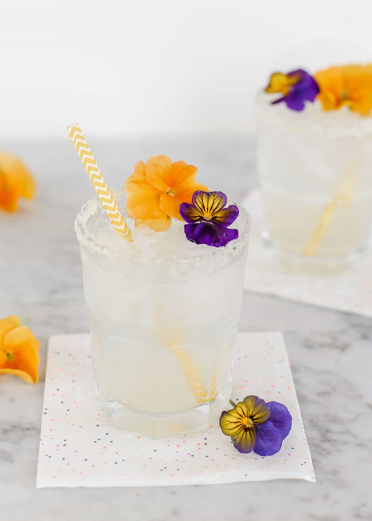two margaritas in short glasses with salt rim and flower garnishes.