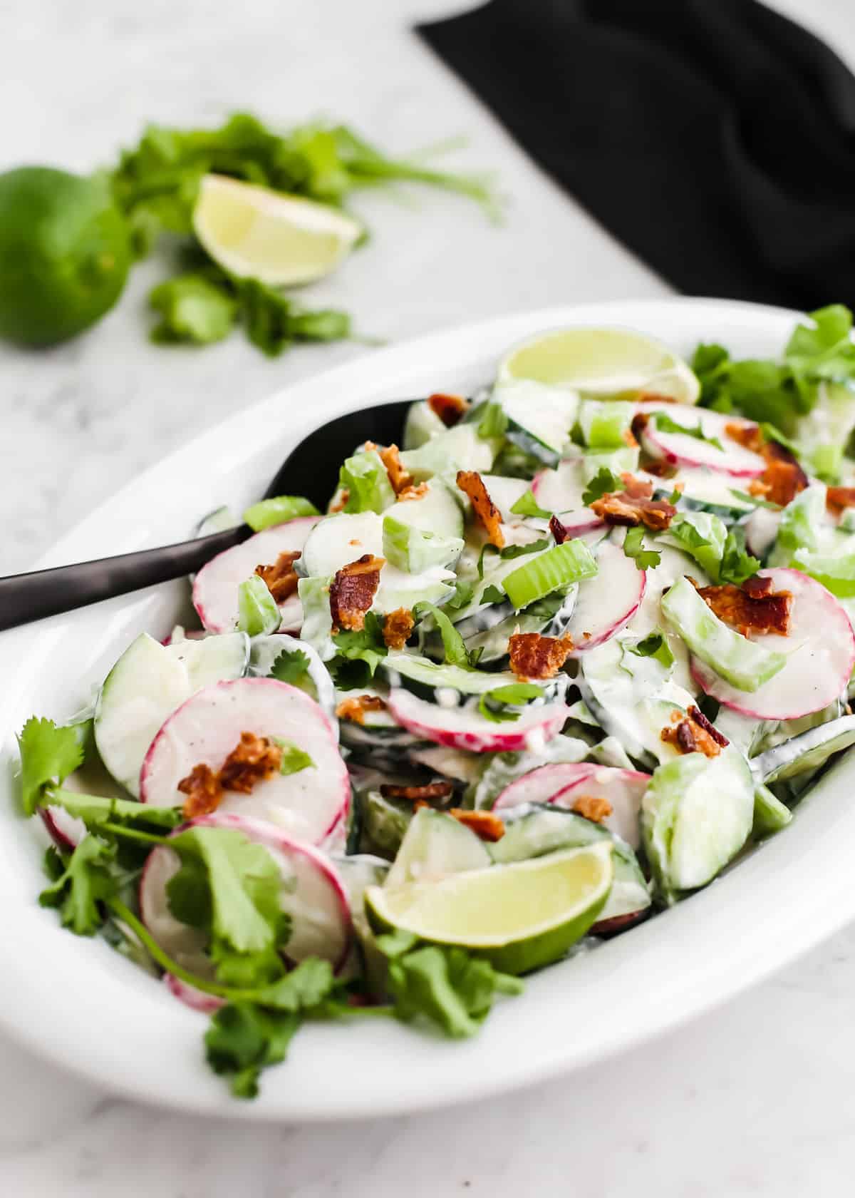 cucumber salad with bacon and radishes and limes, in white oval platter on white marble table.