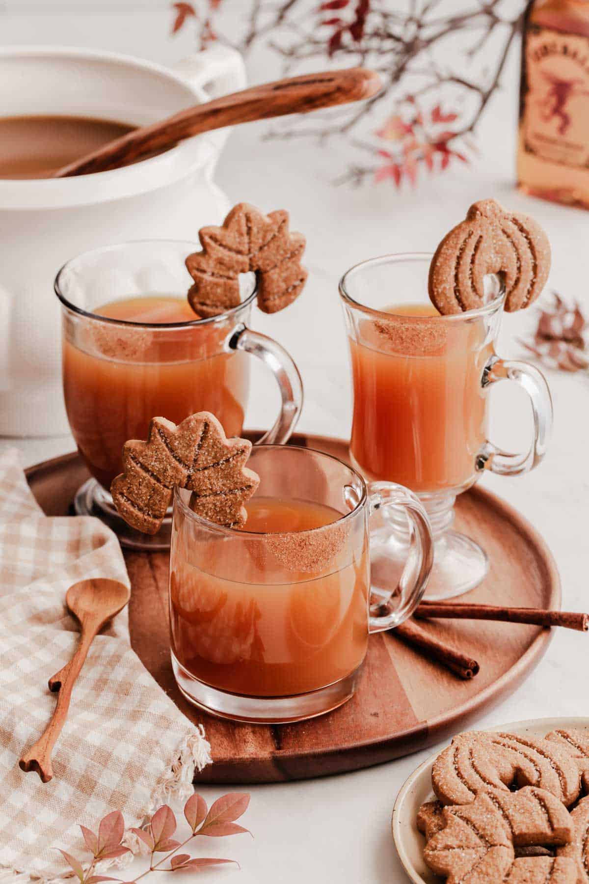 round tray with 3 glass mugs filled with hot cider and cookie garnish on the rim of mugs.