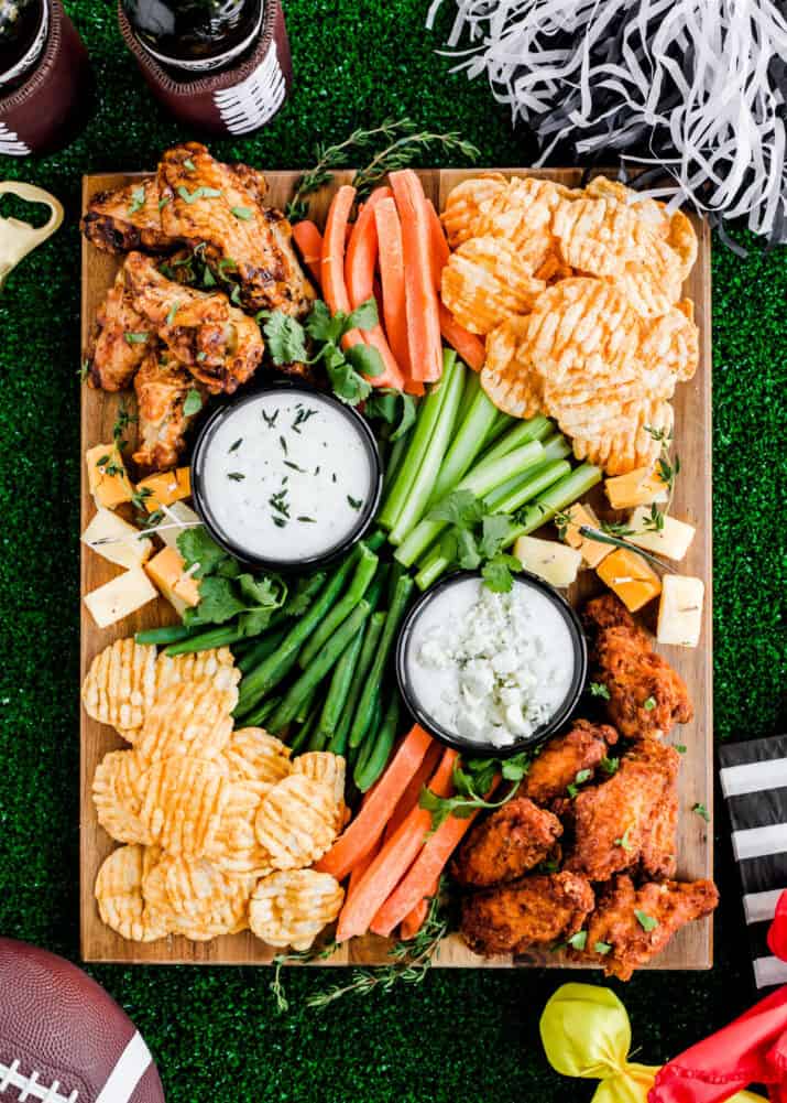 chicken wings, vegetable sticks, white dips, and cheese cubes on wood board on green turf grass surrounded by football party decorations.