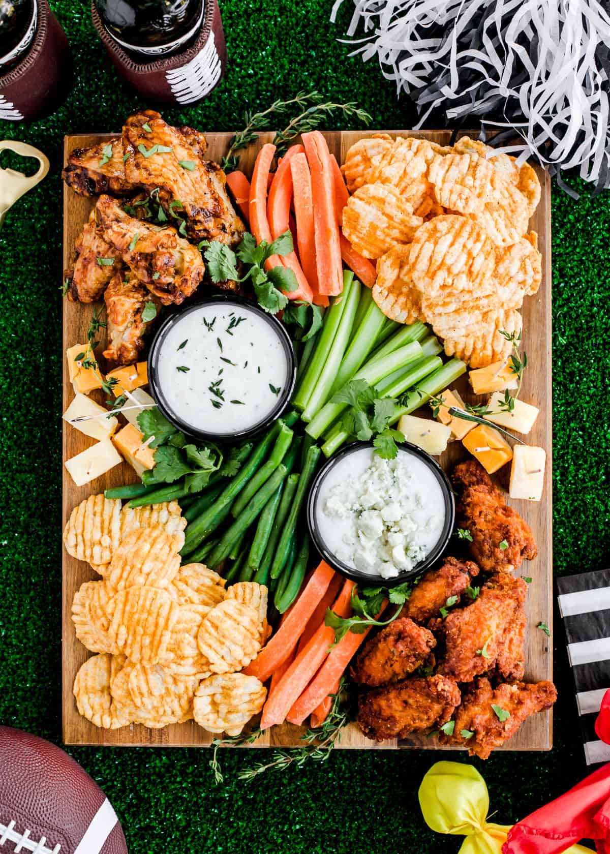 chicken wings, vegetable sticks, white dips, and cheese cubes on wood board, with turf grass background.