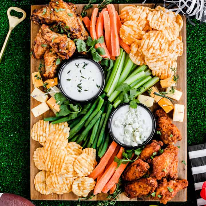 chicken wings, vegetable sticks, white dips, and cheese cubes on wood board.