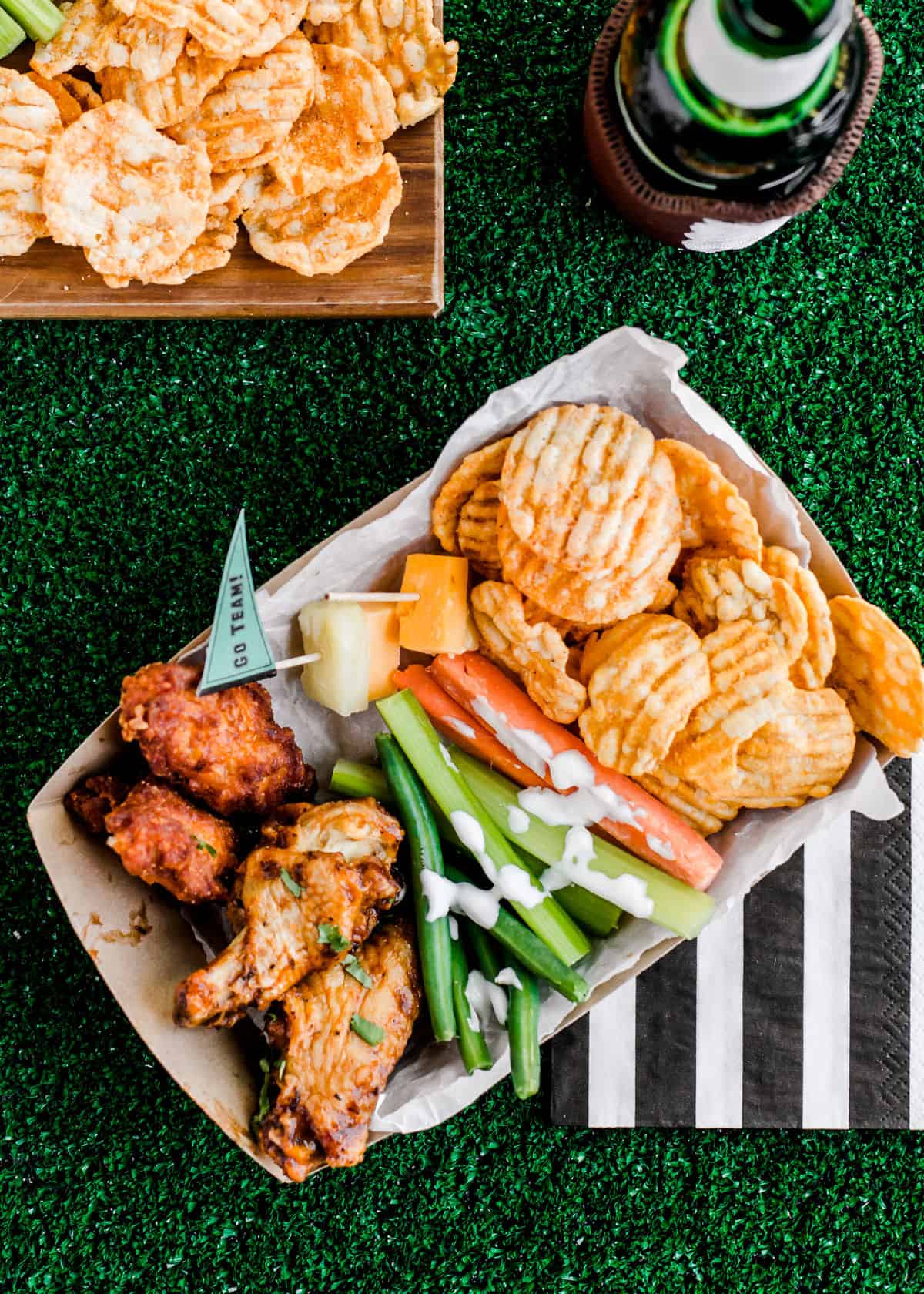 paper snack tray filled with vegetable sticks, chicken wings, and potato chips, on green turf grass background.