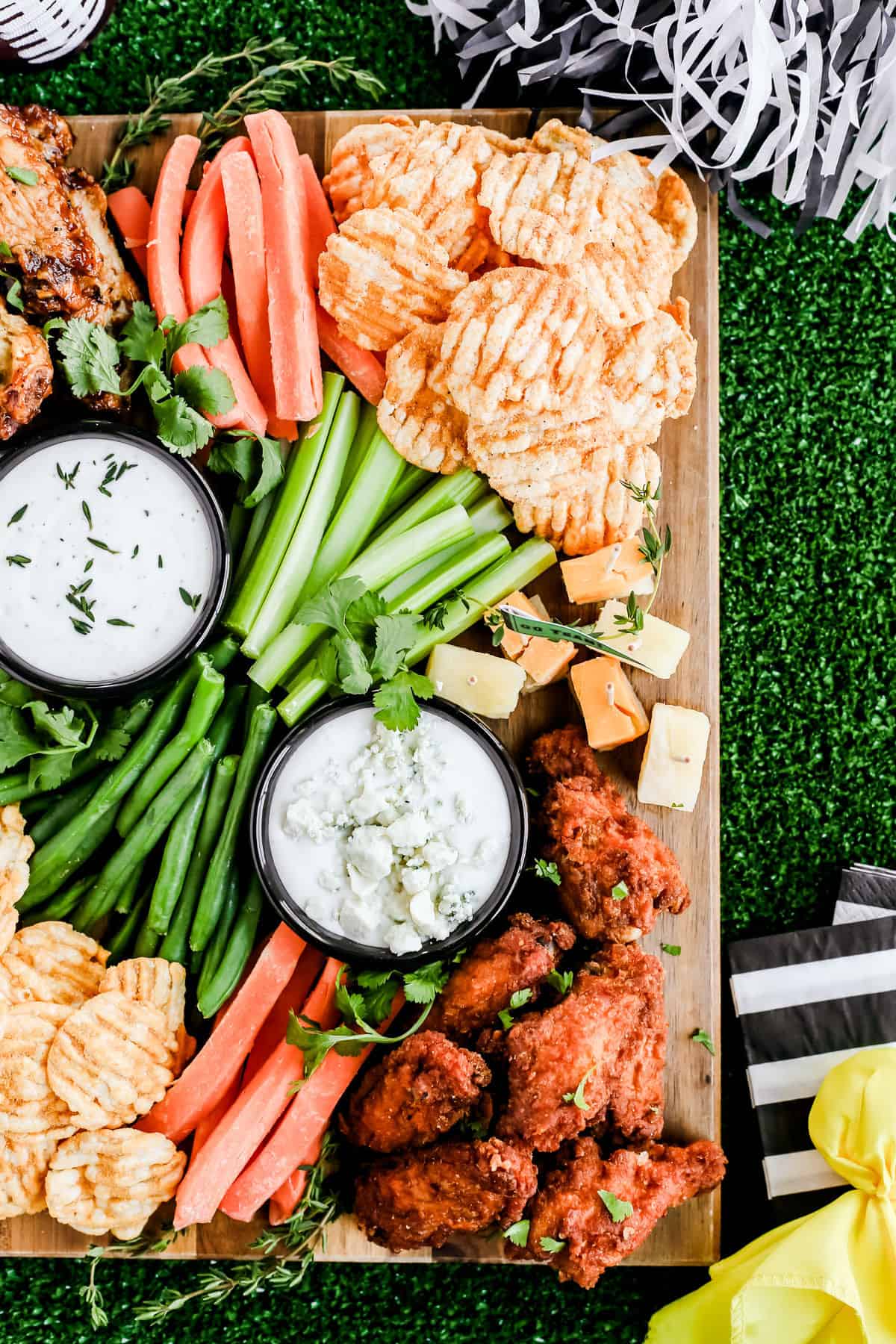 chicken wings, vegetable sticks, white dips, and cheese cubes on wood board surrounded by football themed decorations.