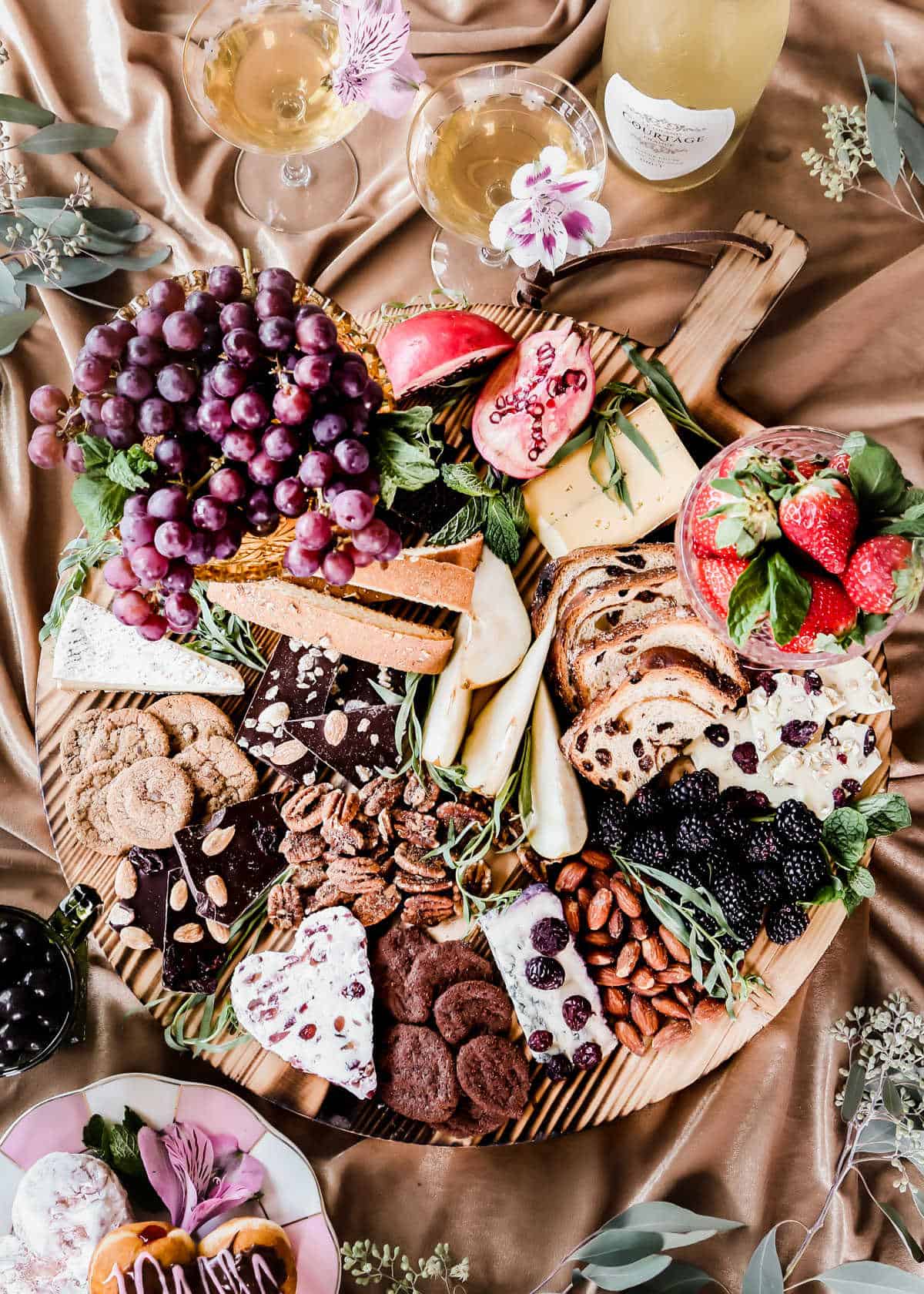 round wood board covered with assortment of chocolates, cheeses, fruit, cookies, and nuts.