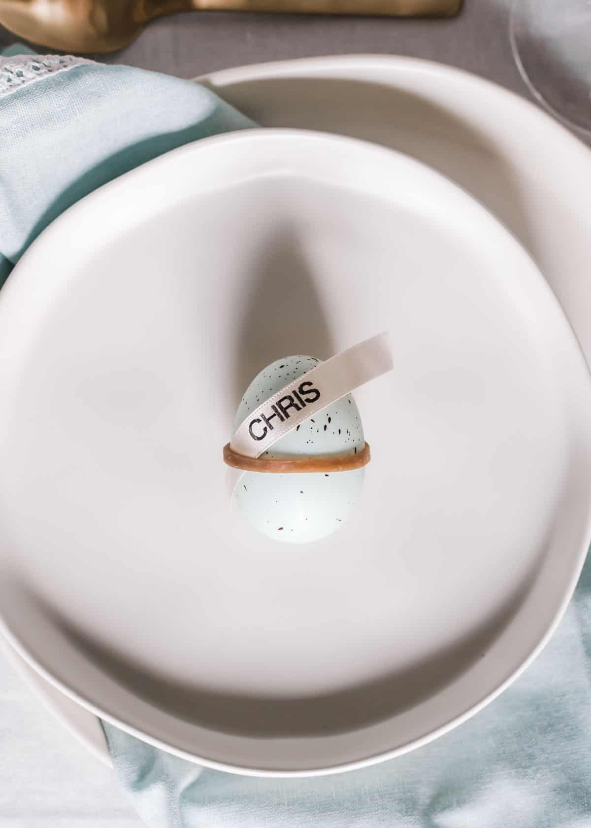 simple egg place card with rubber band around Easter egg and ribbon with name on it tucked into band, on plate.
