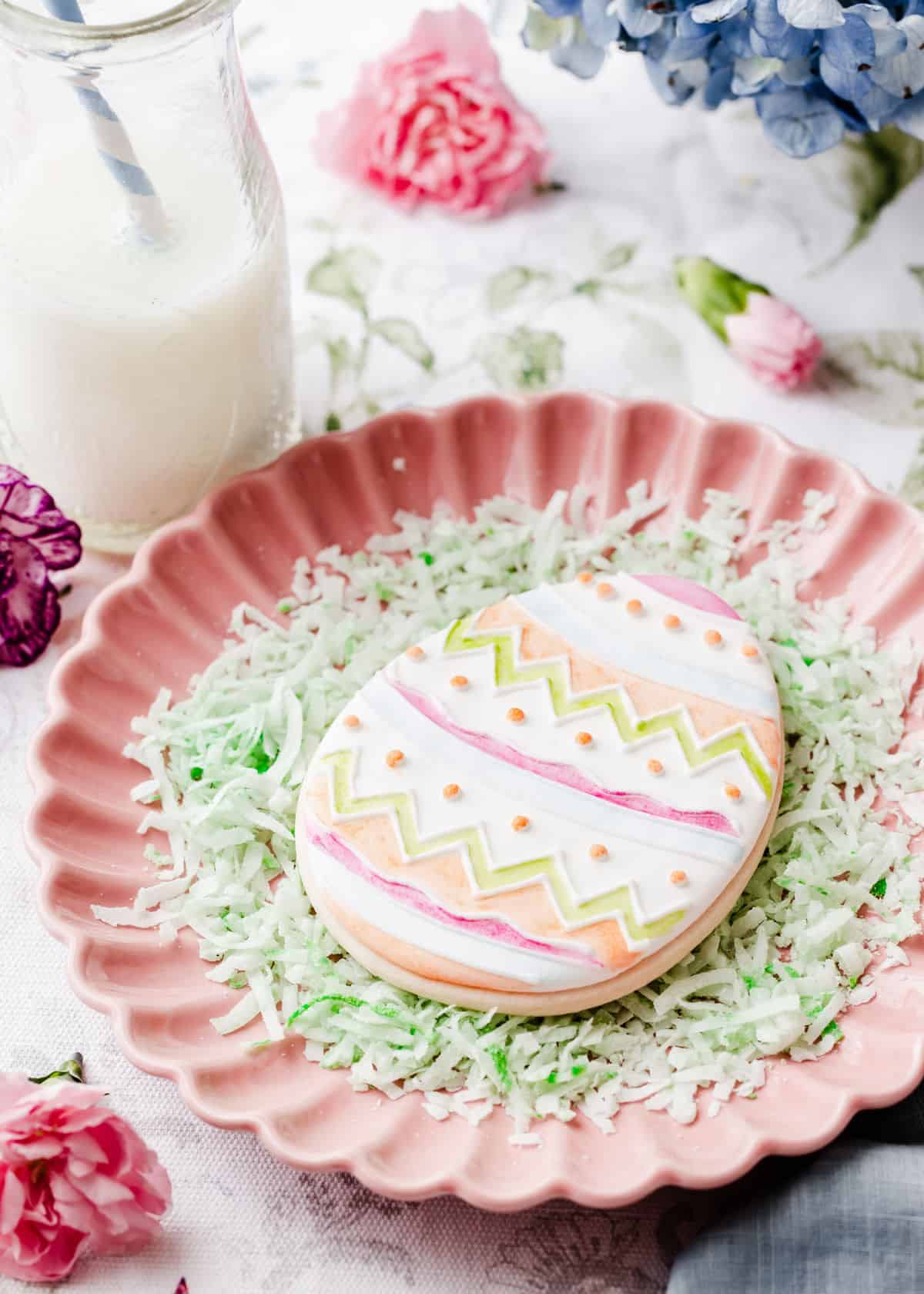 egg shaped cookie with white icing and painted with edible cookie coloring, on pink plate.