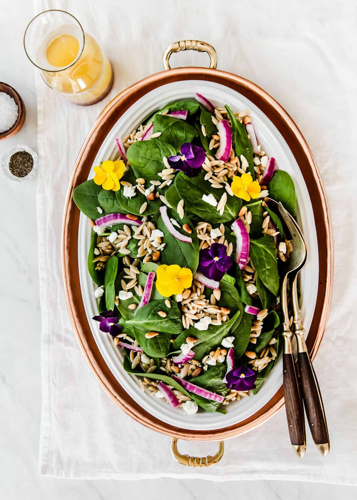 spinach salad in oval platter, with orzo and red onion and dressing on the side, garnished with fresh flowers.