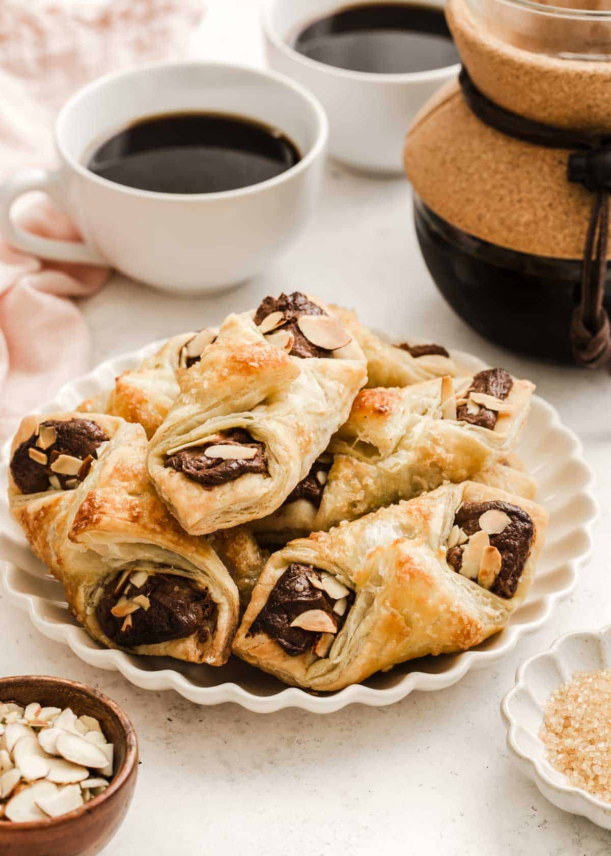 puff pastry packets with chocolate and almonds piled on a scalloped plate with coffee cups in the background.