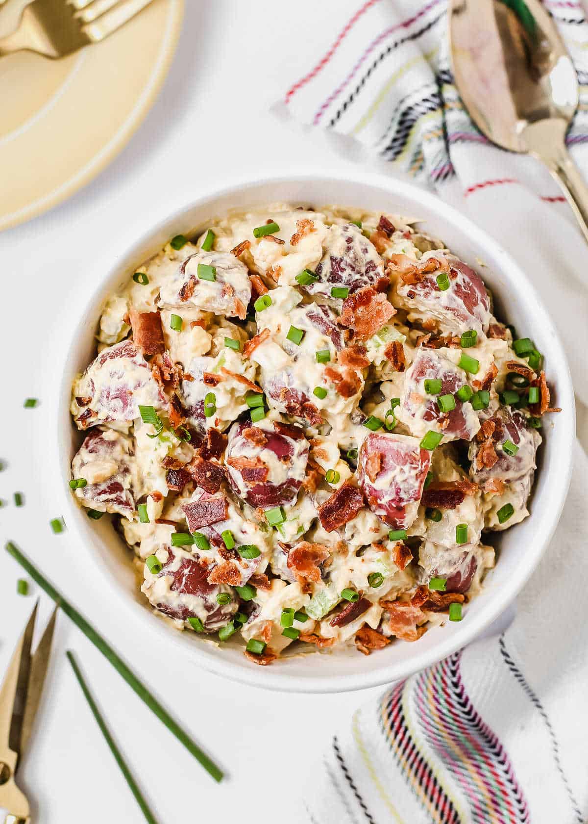 classic potato salad in white bowl surrounded with napkin, spoon, chopped chives, overhead view.