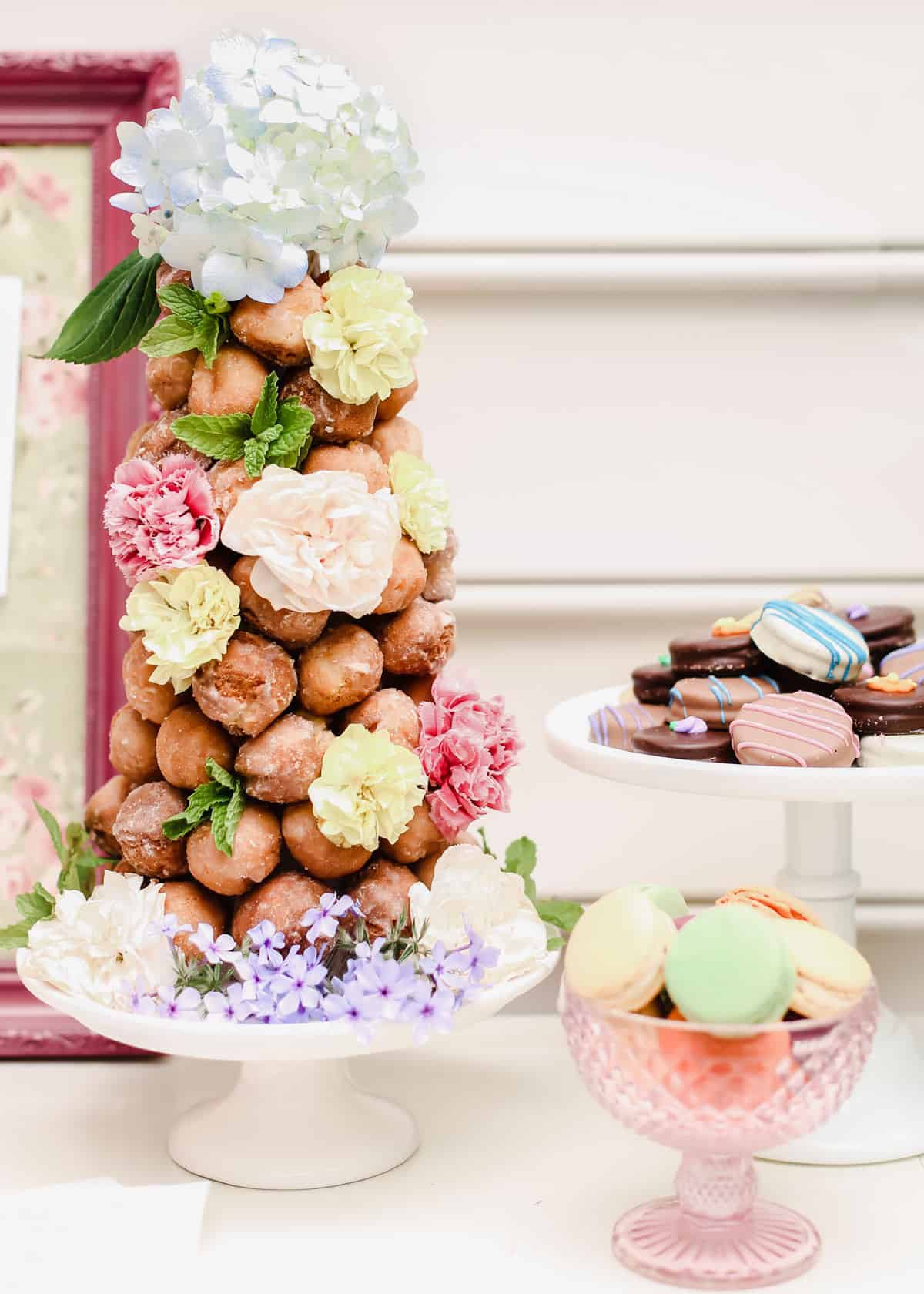 donut hole tree with fresh flowers for garnish, on table beside dish of macarons and chocolates.