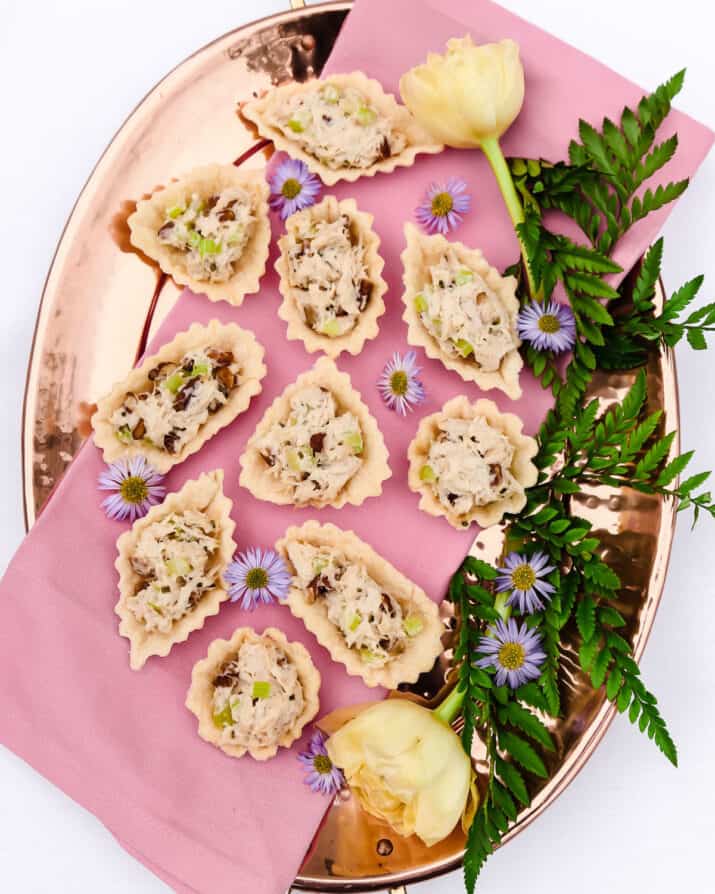 chicken salad in mini pastry cups on pink napkin on copper tray, garnished with flowers and greenery.