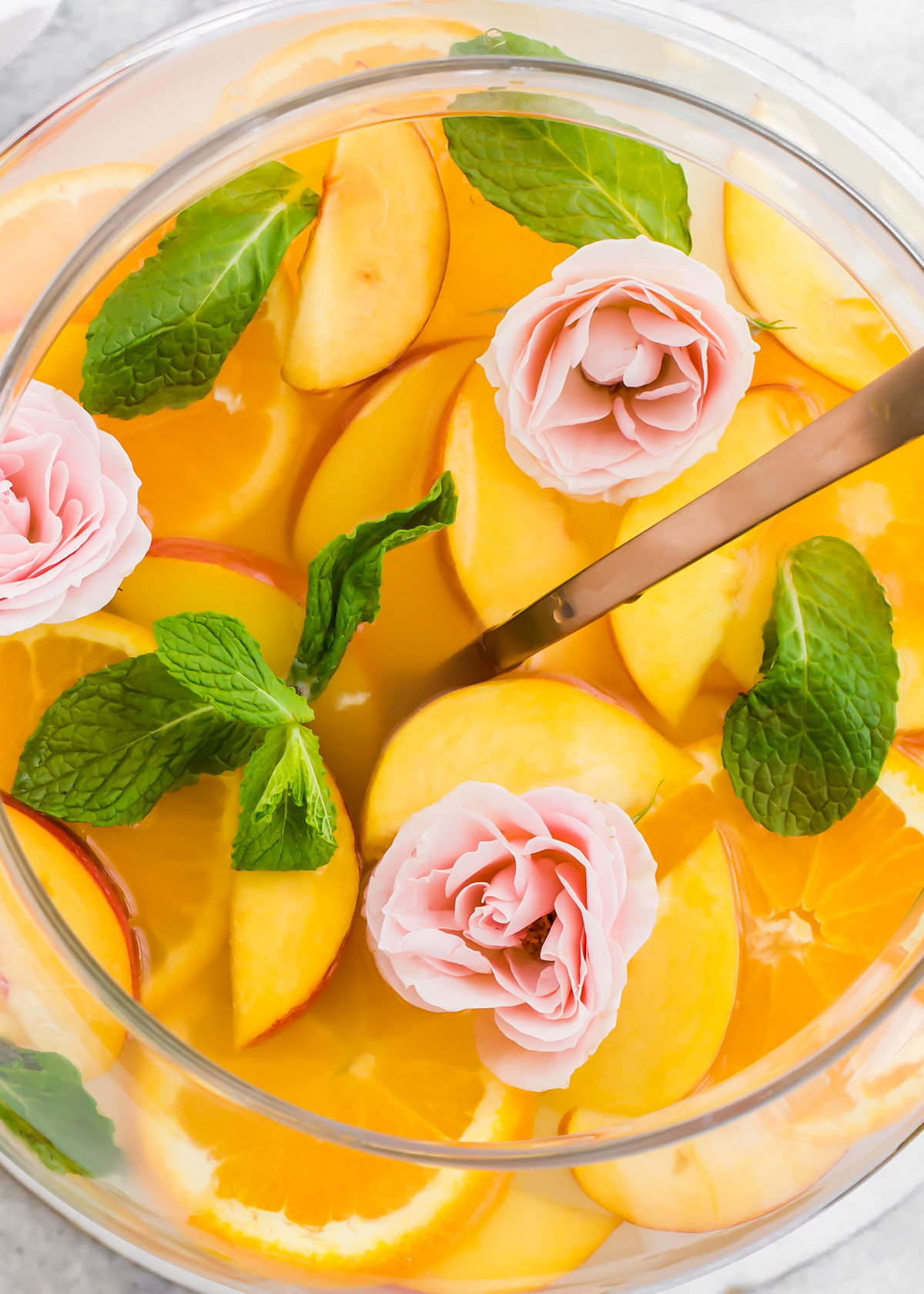 overhead close up of orange colored punch in bowl filled with peach slices, orange slices, mint leaves and roses floating on top.