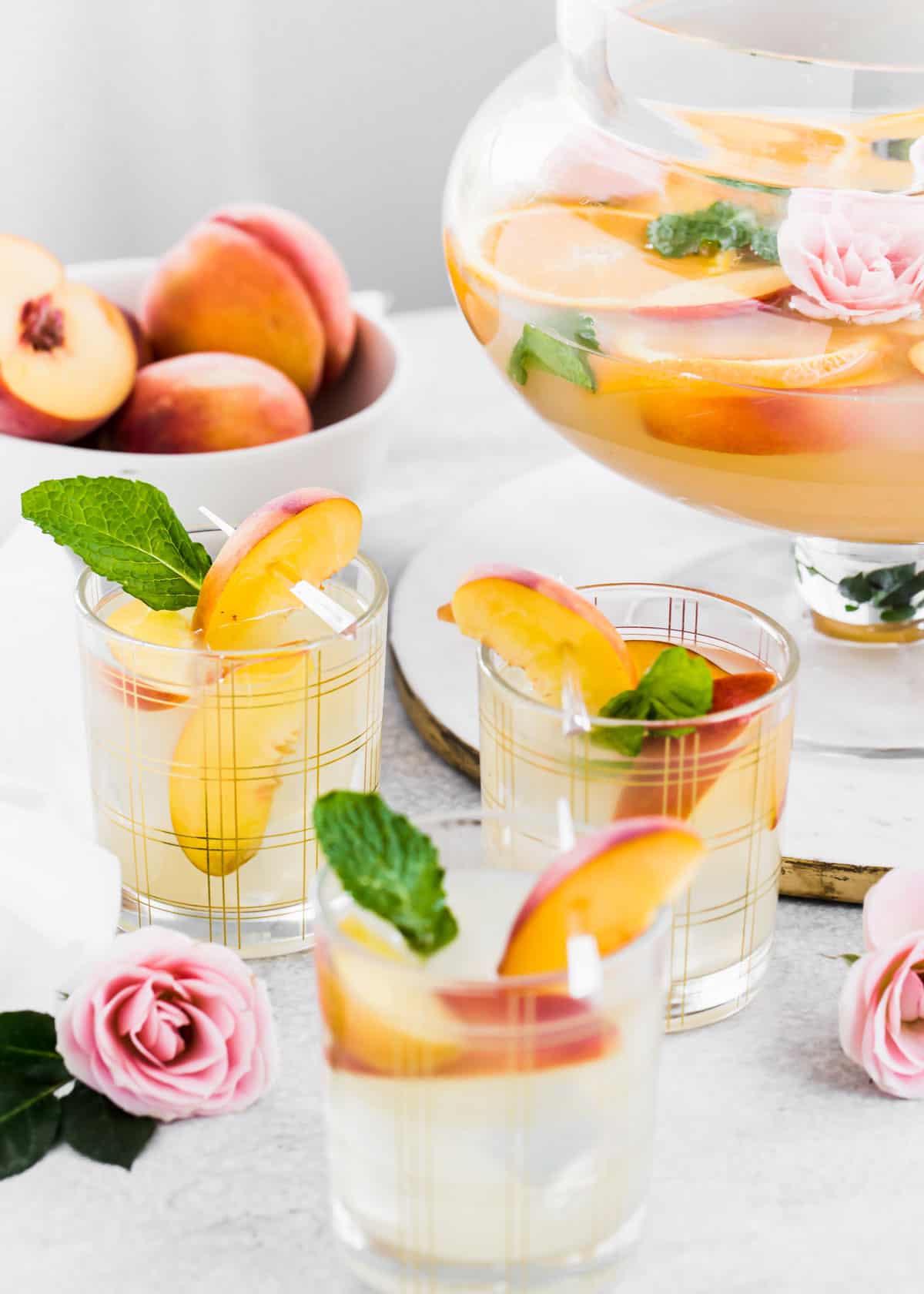 three glasses filled with peach punch and peach slices, with punch bowl and bowl of peaches in background.