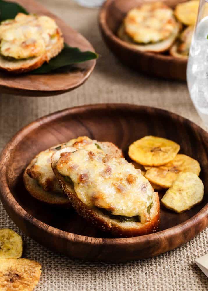 crostini topped with pickle, ham and melted cheese on plate with banana chips.