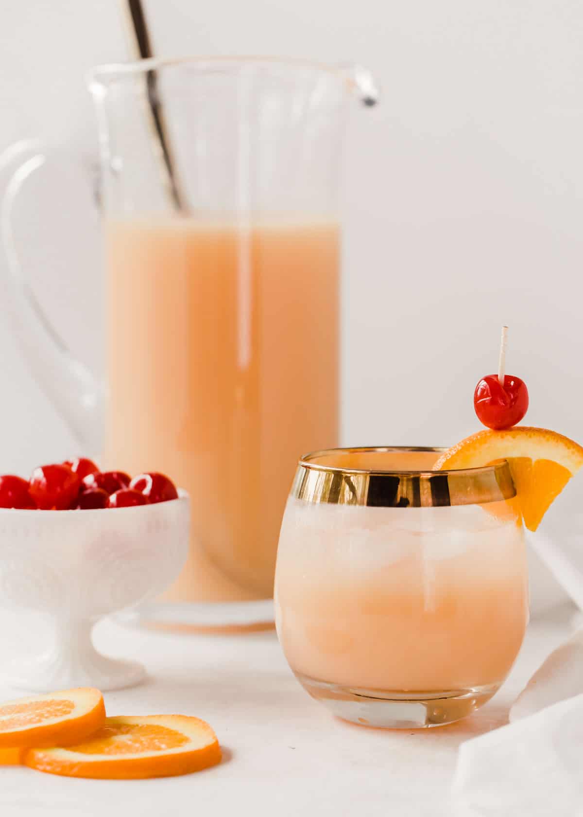 glass of orange drink with orange and cherry garnish, full pitcher in background and dish filled with cherries.