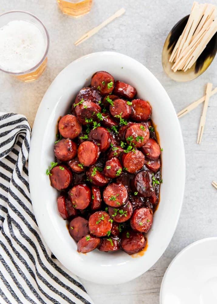 white oval platter with sliced smoked sausage bites in bbq sauce, with picks and drinks on the side.