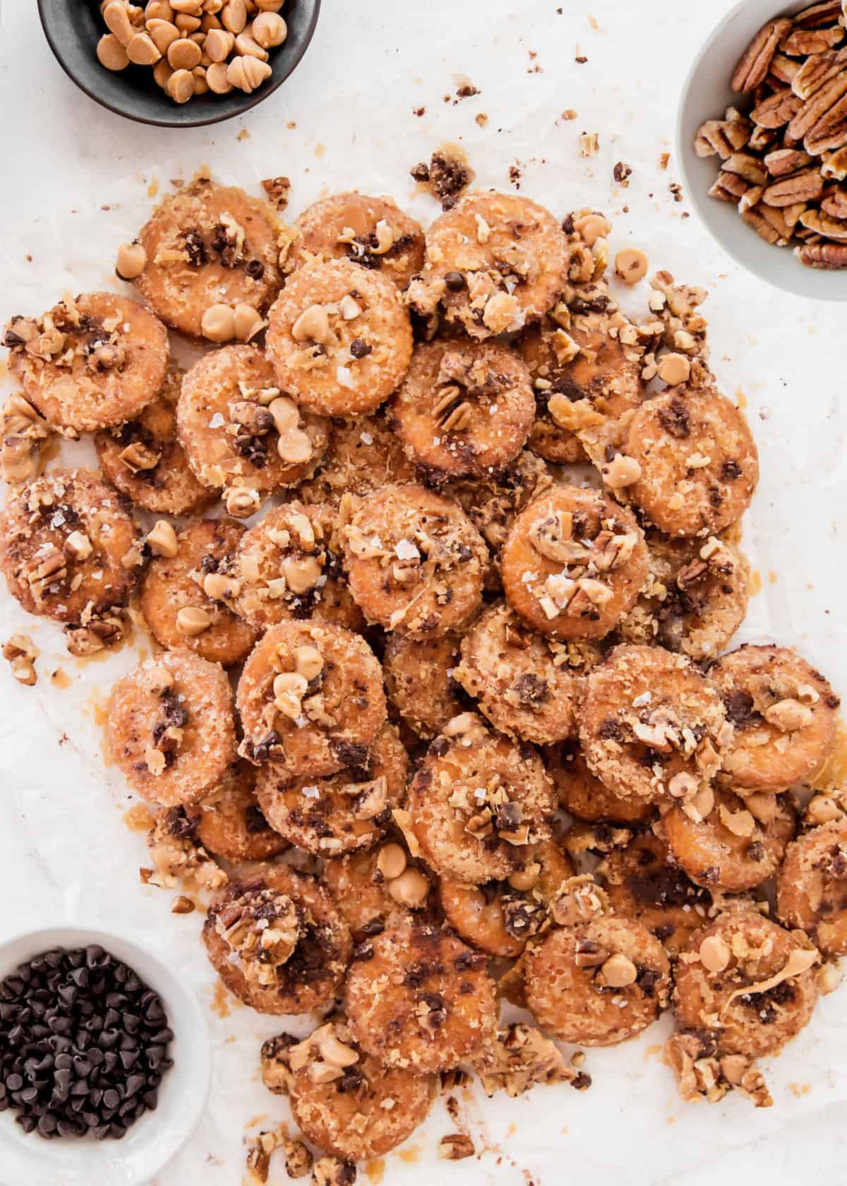 pile of Ritz cracker toffee on white background surrounded by bowls of peanut butter chips, chocolate chips, and pecans.