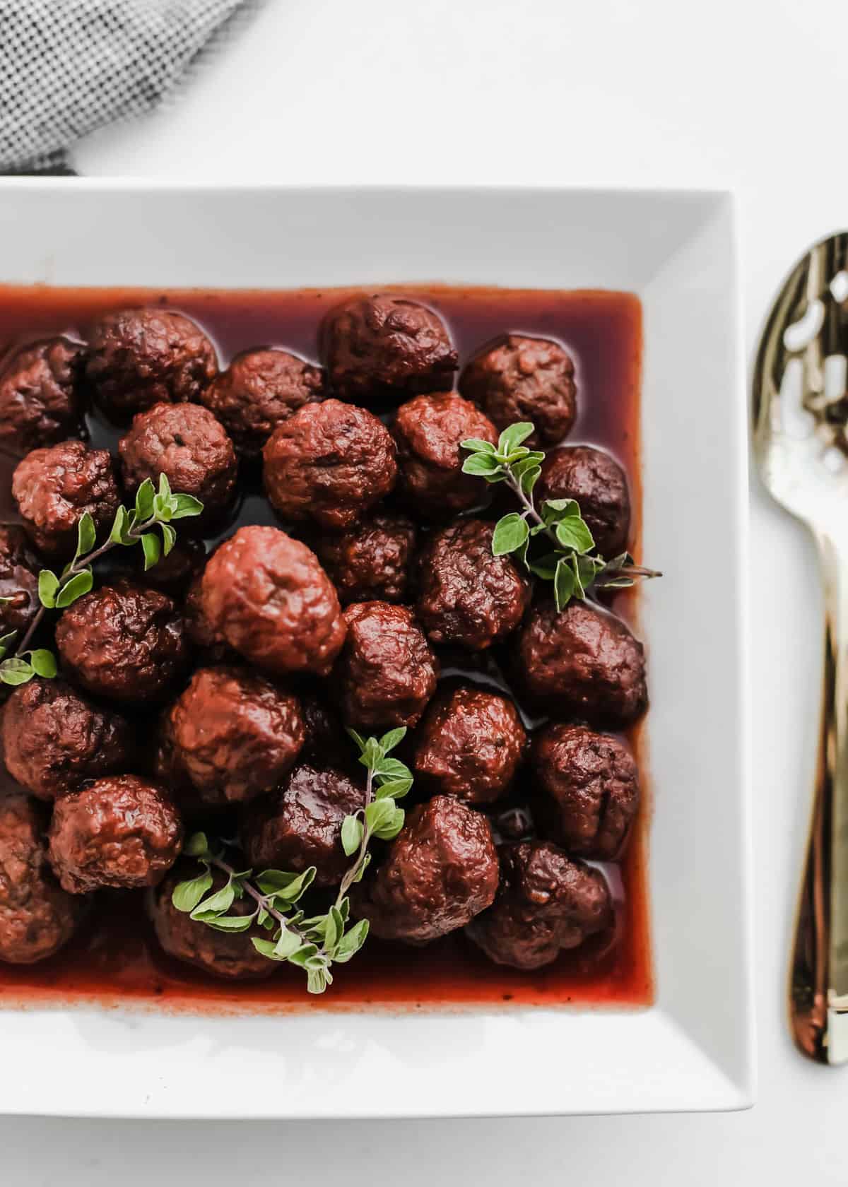 saucy meatballs appetizers piled in a white square dish garnished with herbs, overhead.